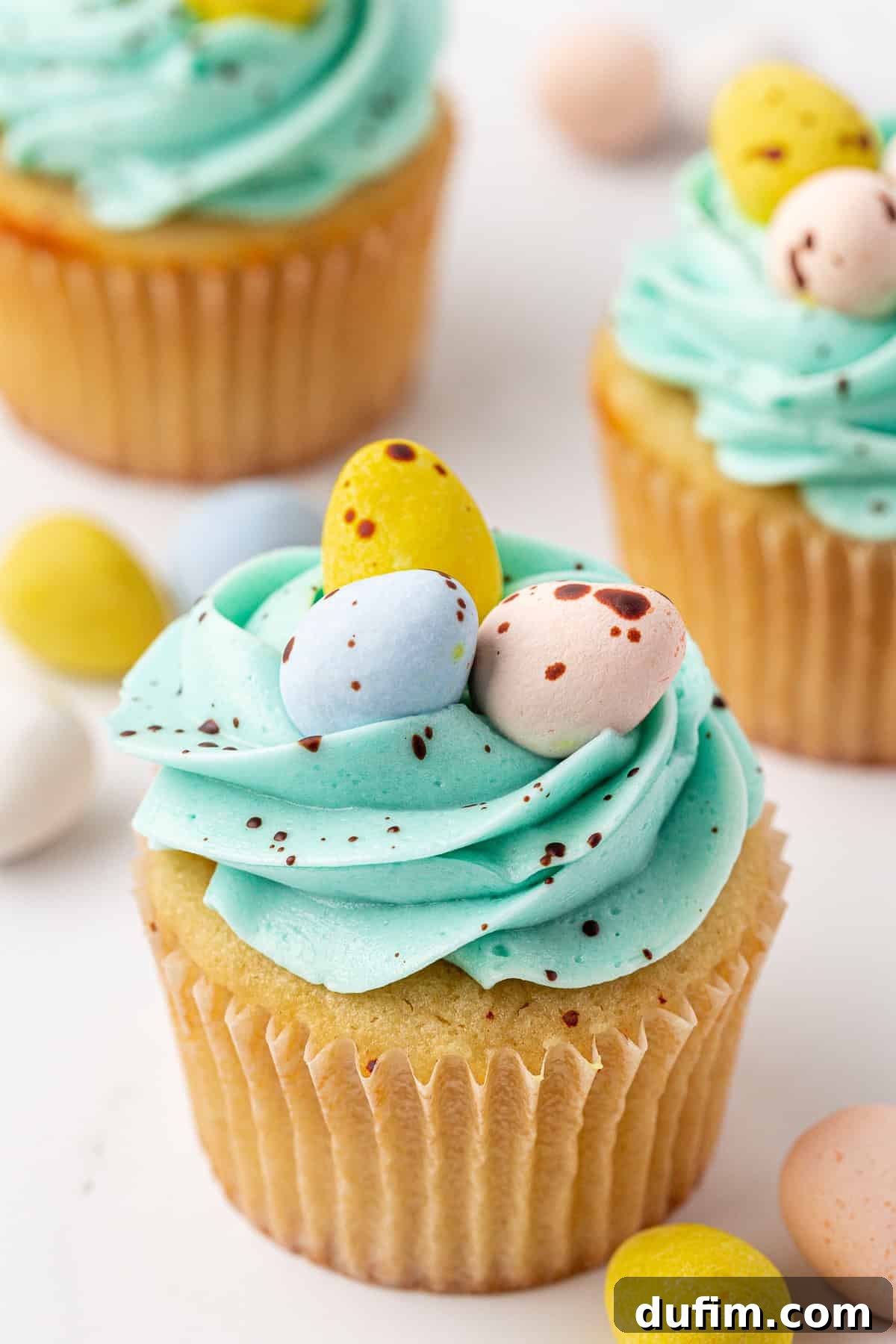 A vanilla cupcake with light blue frosting, topped with three pastel-colored candy eggs, sits on a white surface. Other decorated cupcakes and candy eggs are visible in the background.