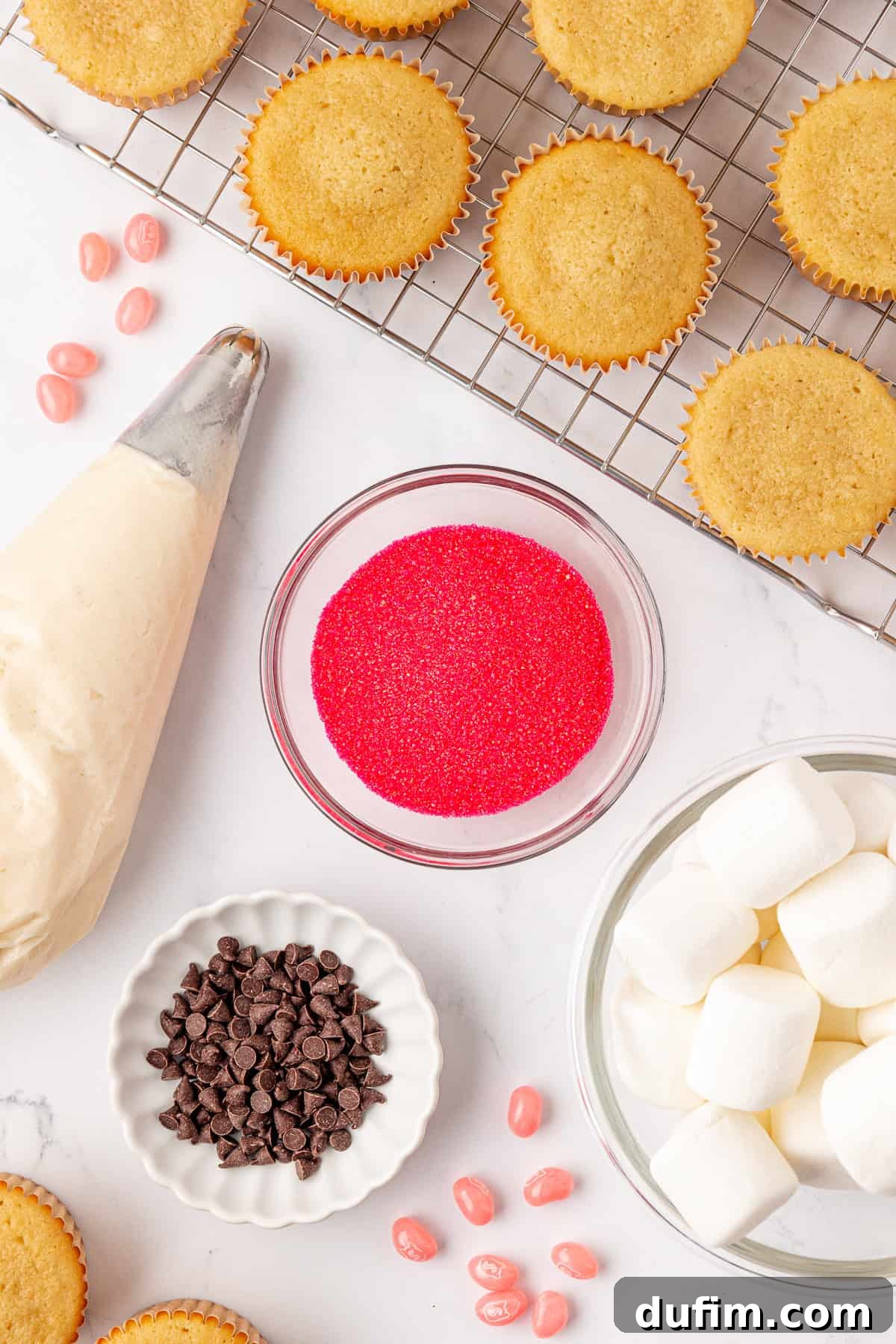 Cupcakes on a cooling rack, with baking supplies nearby: a piping bag filled with frosting, a bowl of red sugar sprinkles, chocolate chips, large marshmallows, and pink candy pieces on a white surface.