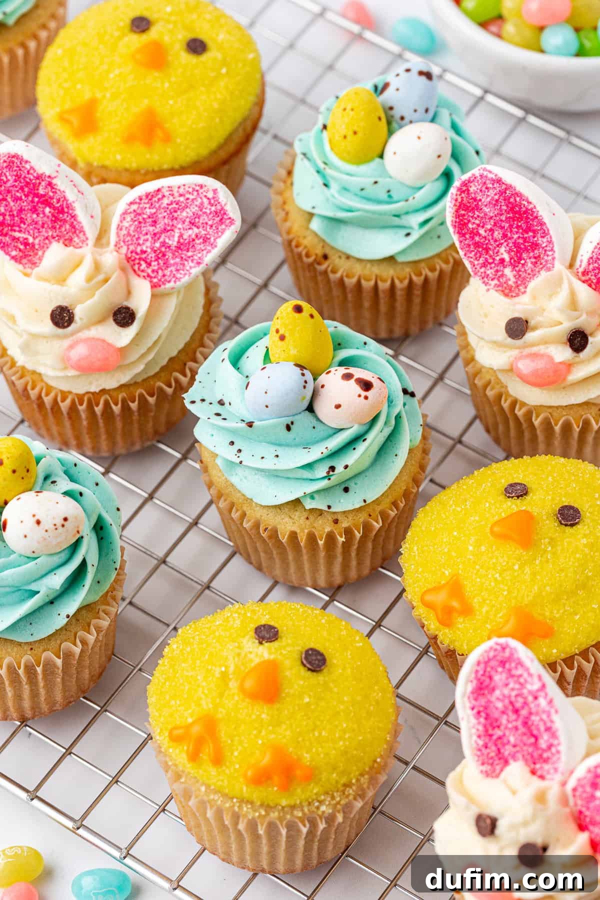 A variety of Easter-themed cupcakes on a cooling rack, decorated as yellow chicks, bunny faces with pink ears, and blue swirled nests topped with candy eggs.