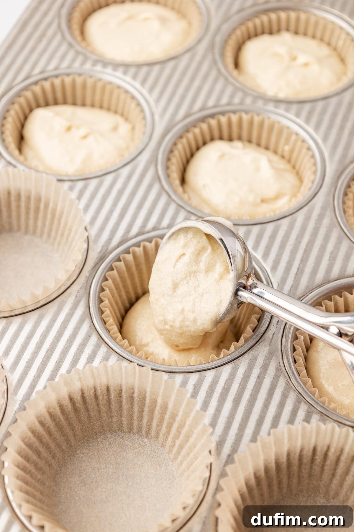 A metal scoop fills a paper-lined muffin tin with batter. Some cups are already filled, while others are empty and ready for batter to be added.