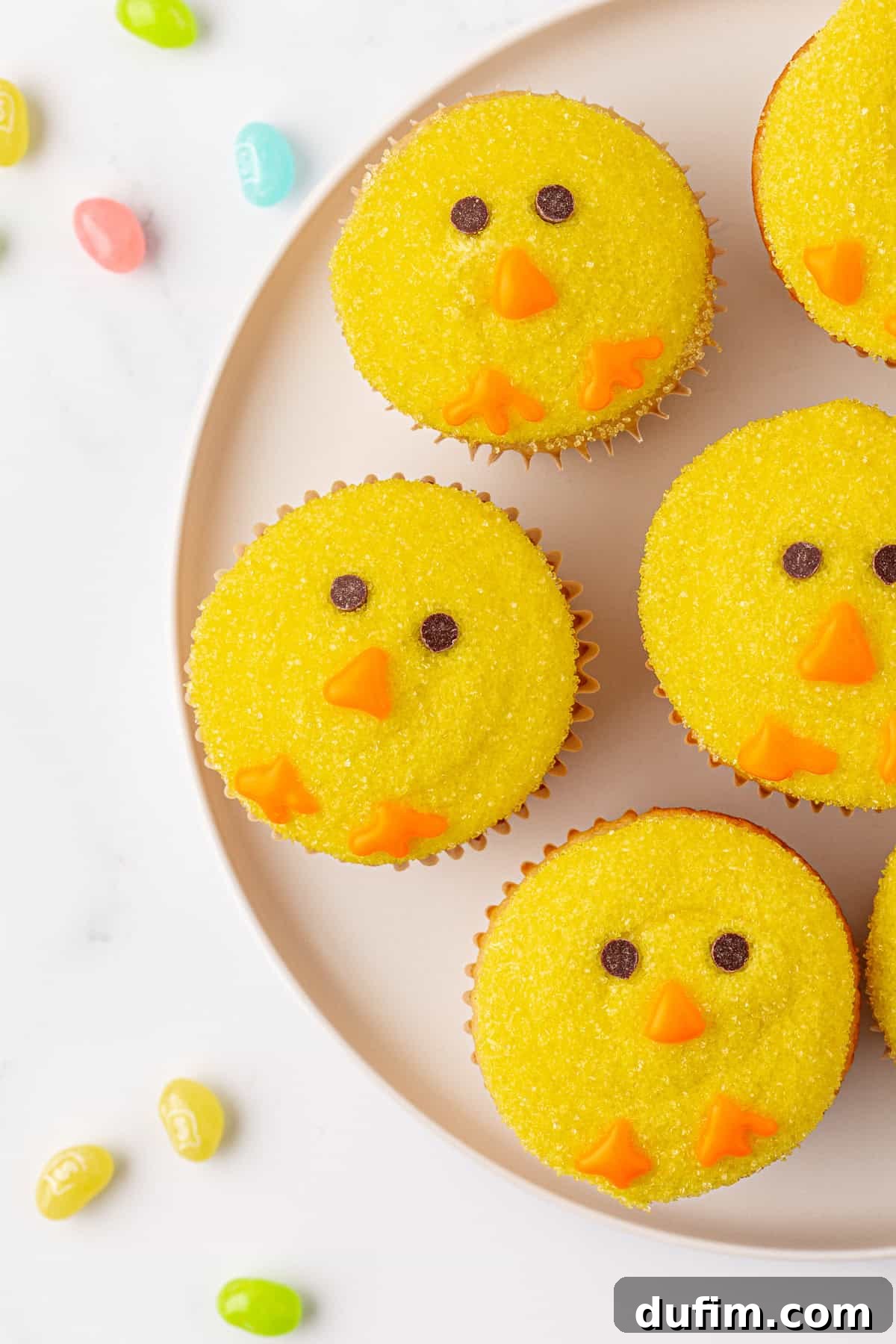 Cupcakes decorated to look like yellow chicks with orange candy beaks and feet, and chocolate eyes, arranged on a round plate. Colorful jelly beans are scattered on the white surface around the plate.