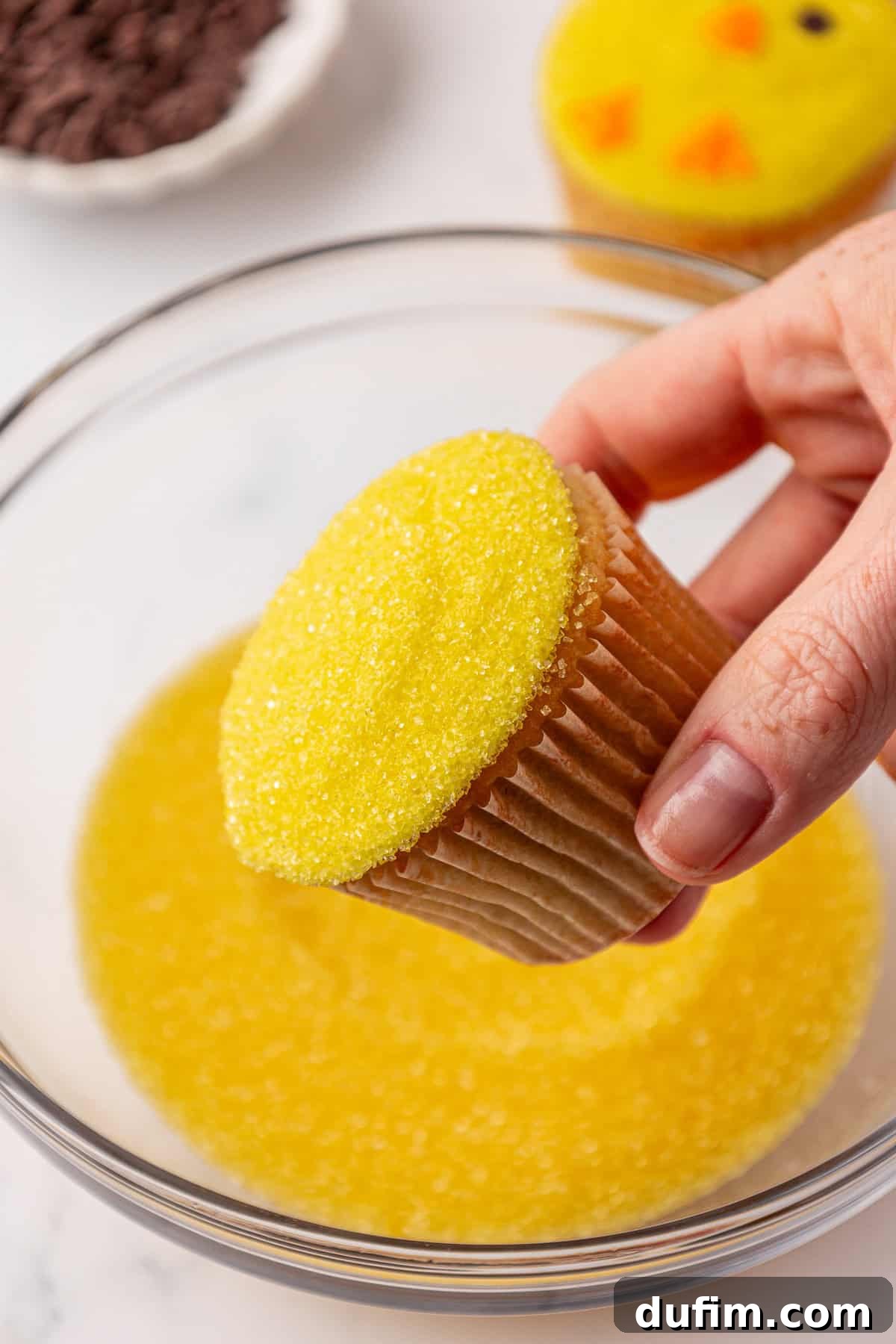 A hand holds a cupcake with yellow sugar sprinkles over a bowl containing more yellow sugar. Another decorated cupcake and a bowl of chocolate chips are visible in the background.
