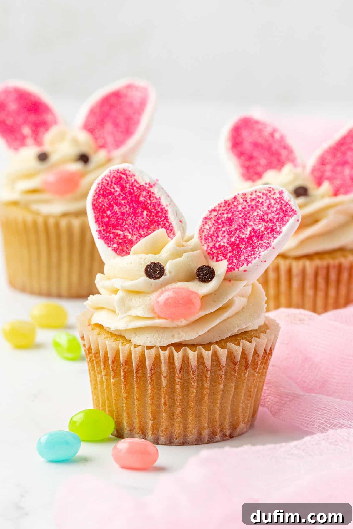 Three cupcakes decorated as bunnies with pink and white candy ears, chocolate chip eyes, and a pink candy nose, surrounded by colorful jellybeans on a white surface with a pink cloth.