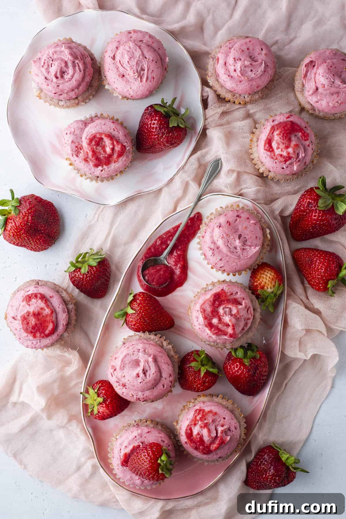 An overhead view of strawberry cupcakes with pink frosting on decorative plates, surrounded by fresh strawberries and a pink cloth, with a spoon holding strawberry sauce.