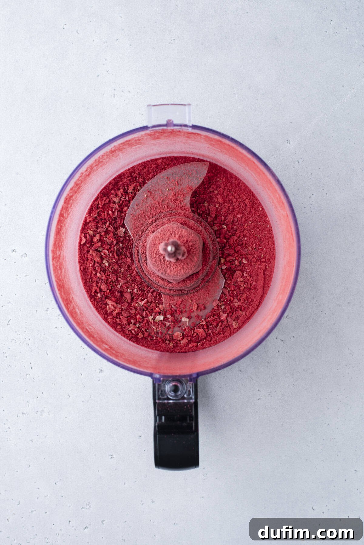 A food processor with bright red powder inside sits on a light gray surface, viewed from above. The lid is off, revealing the metal blade and finely ground contents.