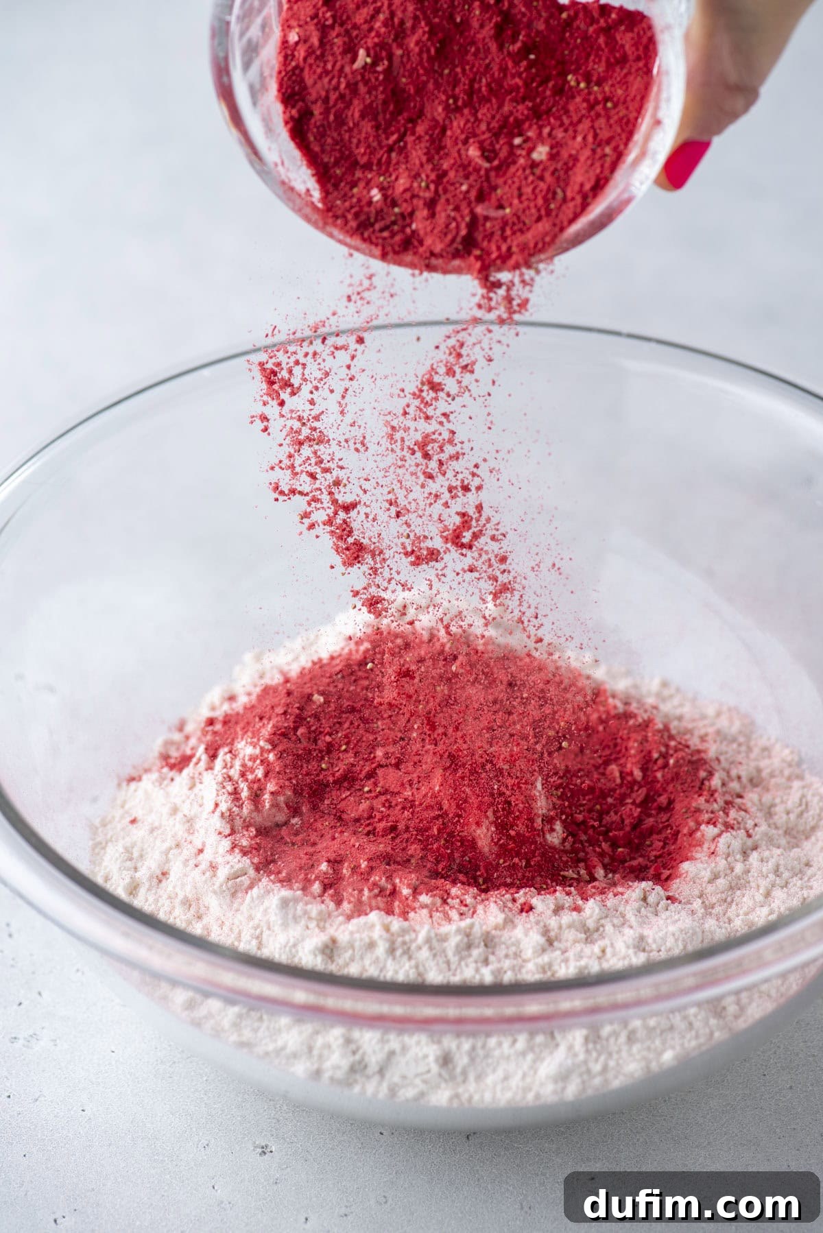 A hand pours vibrant red powder into a clear glass bowl filled with flour, creating a colorful dusting above the mixture on a light gray surface.