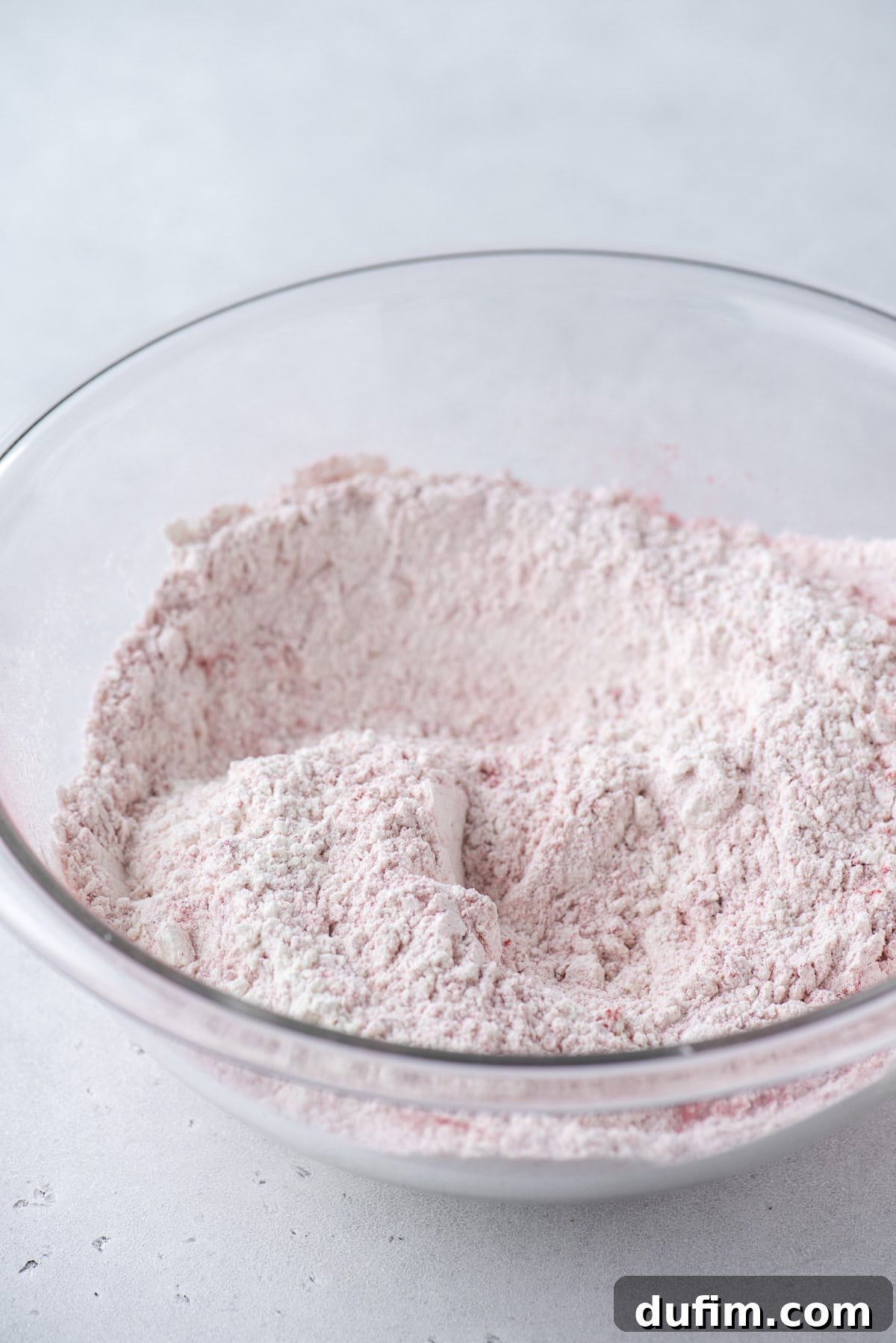 A large clear glass bowl containing a mixture of pale pink dry ingredients, likely flour and other powders, sits on a light gray surface.