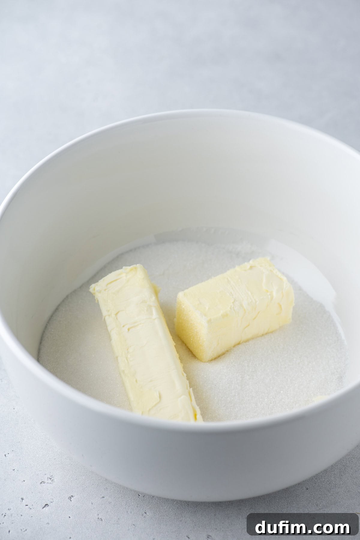 A white bowl contains two sticks of butter and a layer of granulated sugar on a light grey surface.