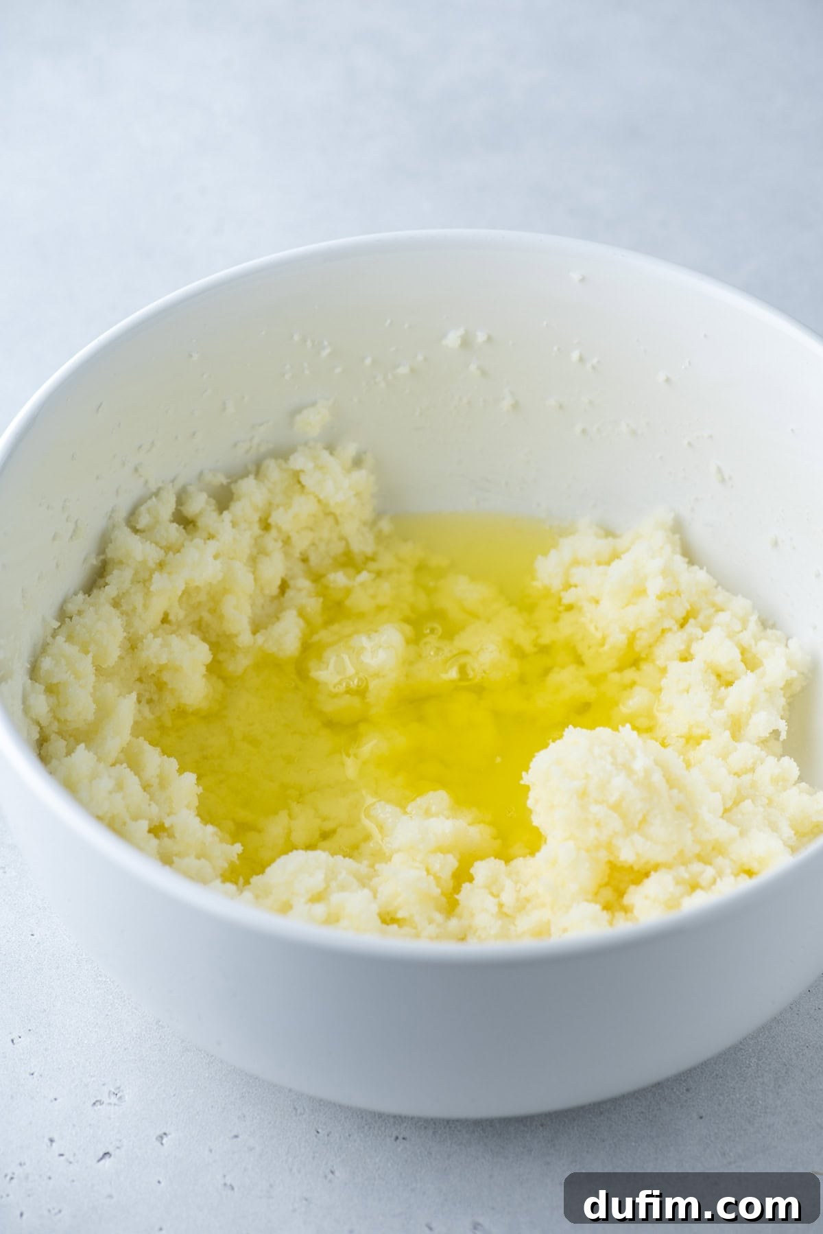 A white bowl containing creamed butter and sugar, with melted butter pooling in the center, sits on a light gray surface.