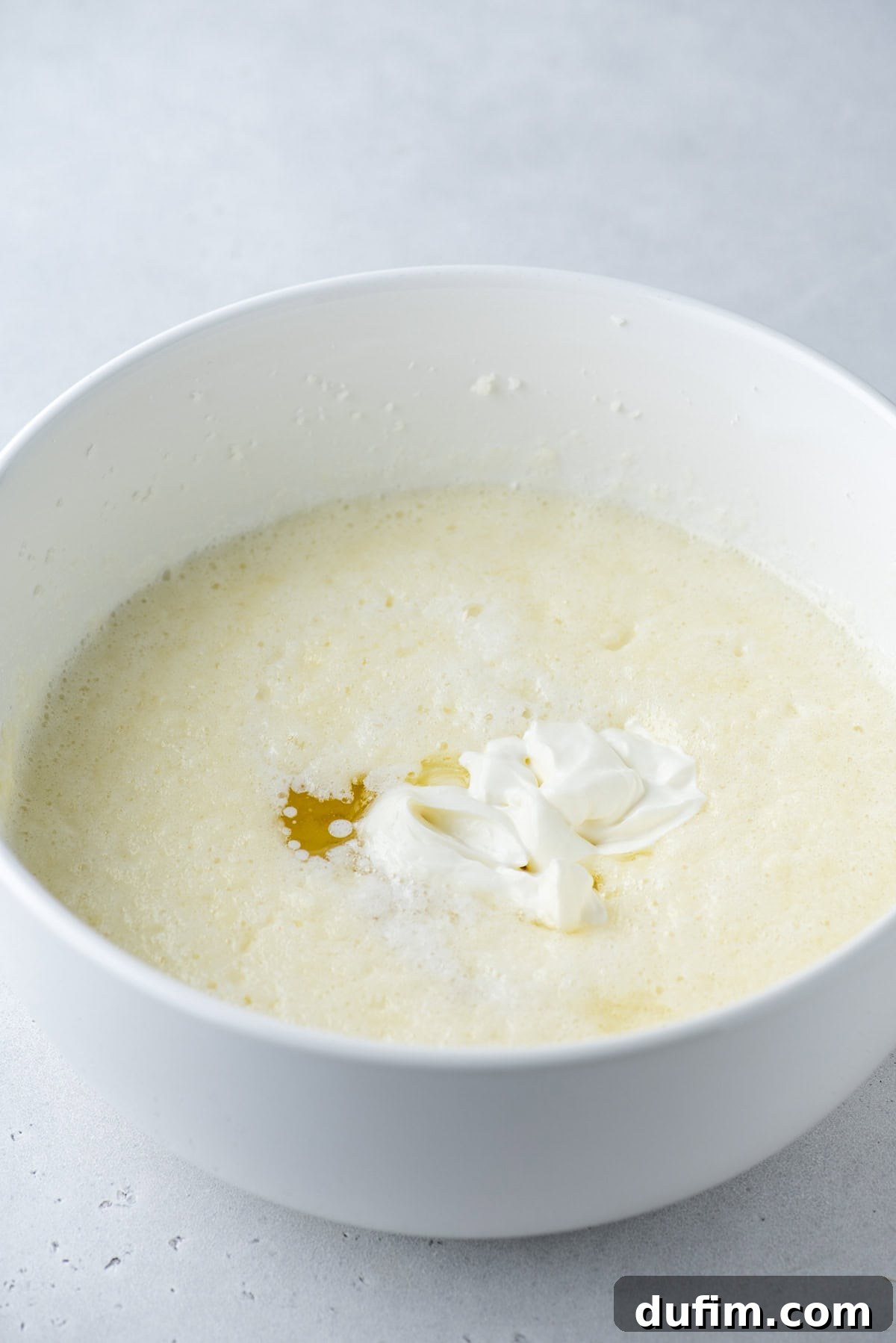 A white mixing bowl filled with a creamy, pale yellow batter, topped with a dollop of sour cream and a small amount of melted butter on a light gray surface.