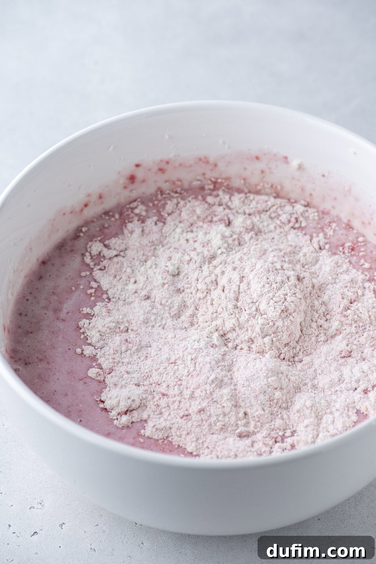 A white mixing bowl filled with pink batter, partially topped with a mound of dry flour mixture, sits on a light gray surface.