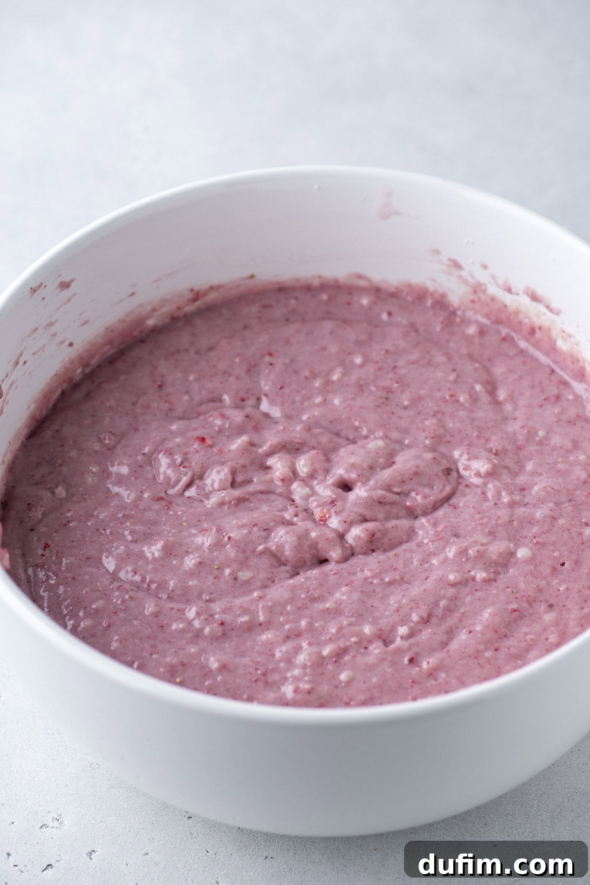 A white mixing bowl filled with thick, pinkish batter, speckled with small bits, sits on a light gray surface.