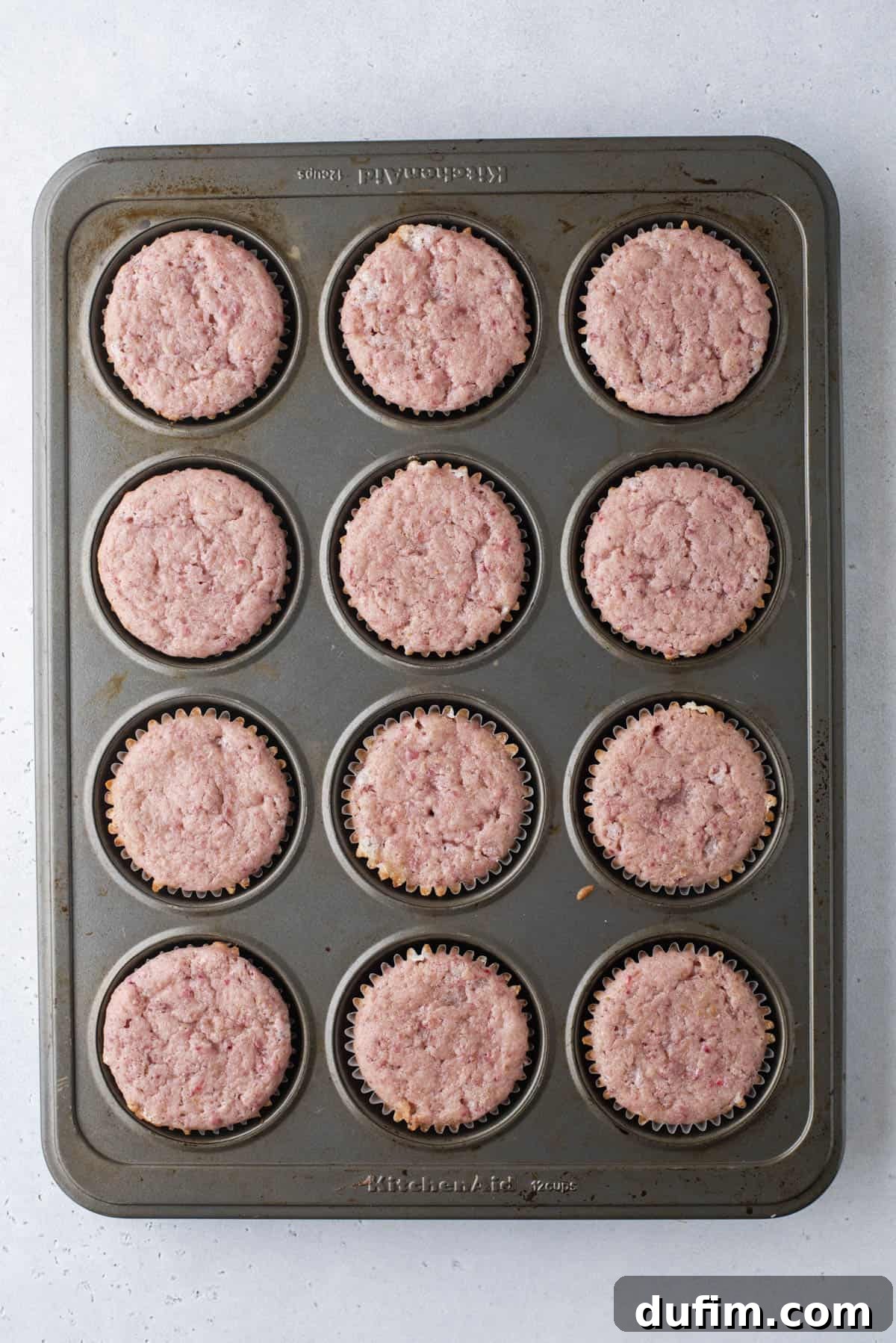 A muffin tin with twelve cupcake liners filled with baked, pinkish muffins, viewed from above on a light gray surface.