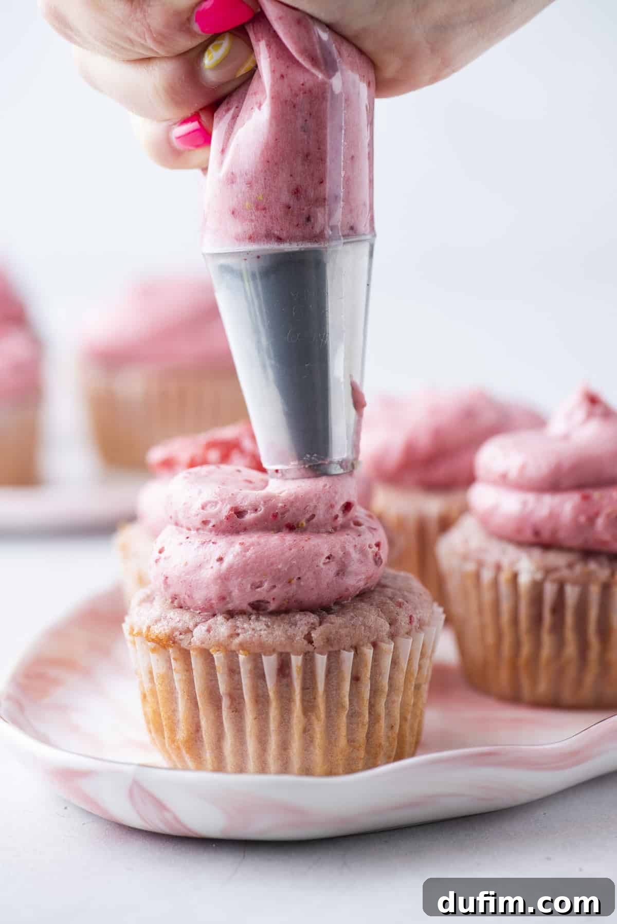 A hand is piping pink strawberry frosting onto a cupcake using a piping bag. Several unfrosted and frosted cupcakes sit on a plate in the background.