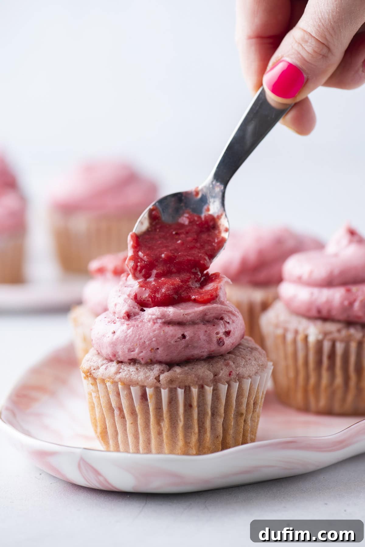A hand with pink nail polish uses a spoon to spread strawberry topping on a cupcake with pink frosting. More similarly decorated cupcakes are visible in the background on a marbled plate.