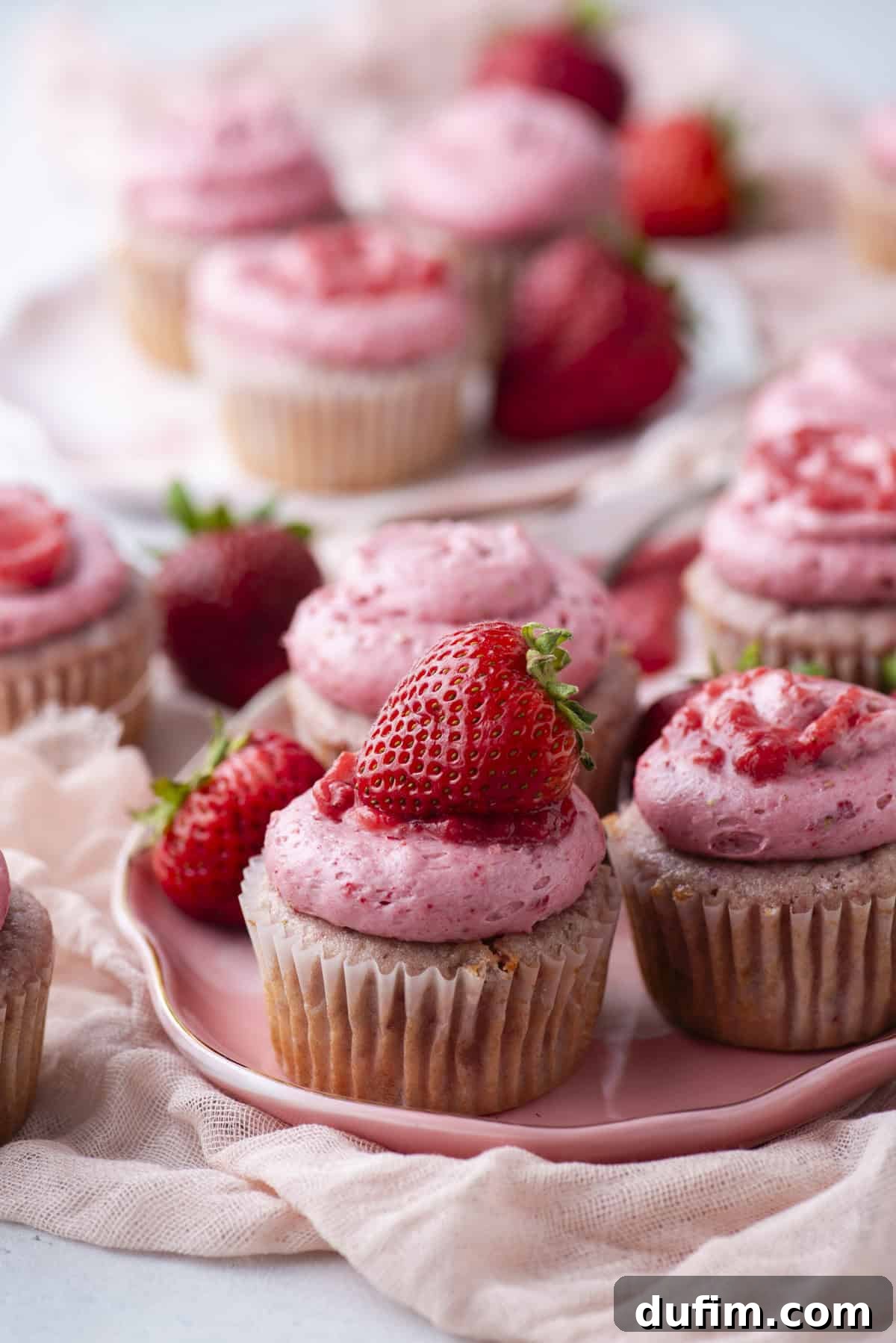 Cupcakes topped with pink strawberry frosting and fresh strawberries are arranged on a pink plate, with more cupcakes and whole strawberries in the background. A soft, light fabric is draped around the plate.