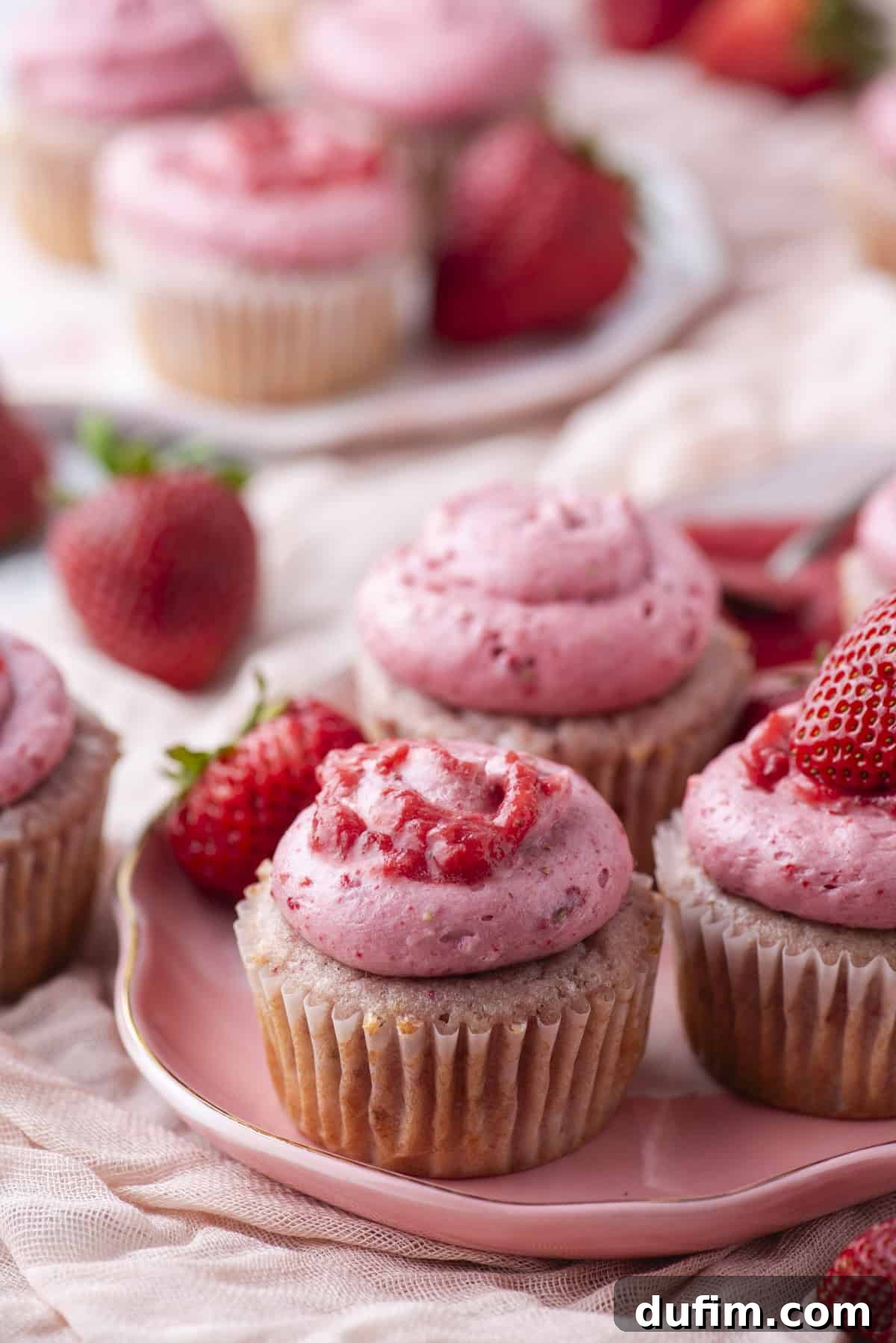 A close-up of vanilla cupcakes topped with pink strawberry frosting and fresh strawberries, arranged on a pink plate with whole strawberries scattered around.