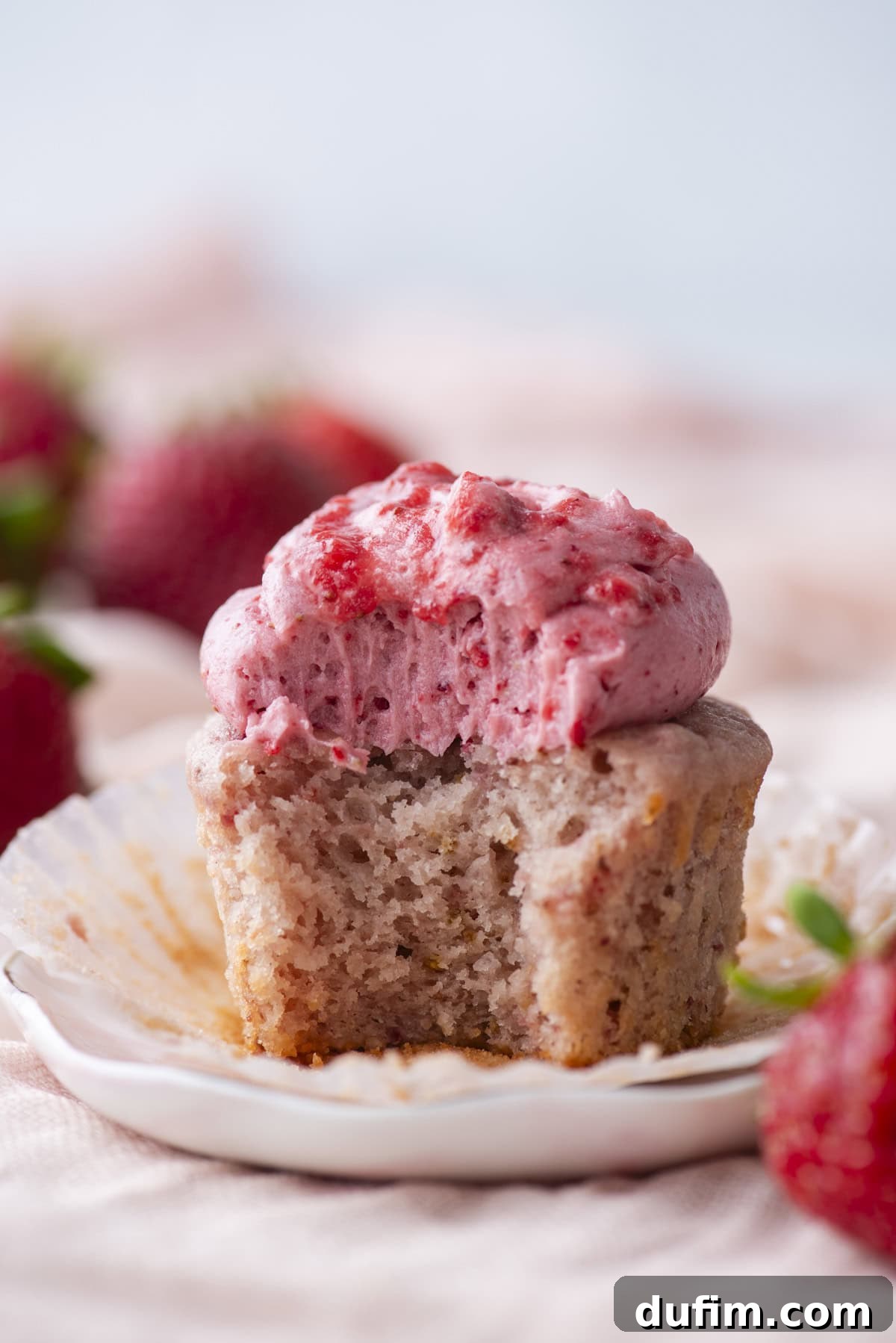 A strawberry cupcake with pink strawberry frosting, partially unwrapped and with a bite taken out, sits on a small plate. Fresh strawberries are blurred in the background.