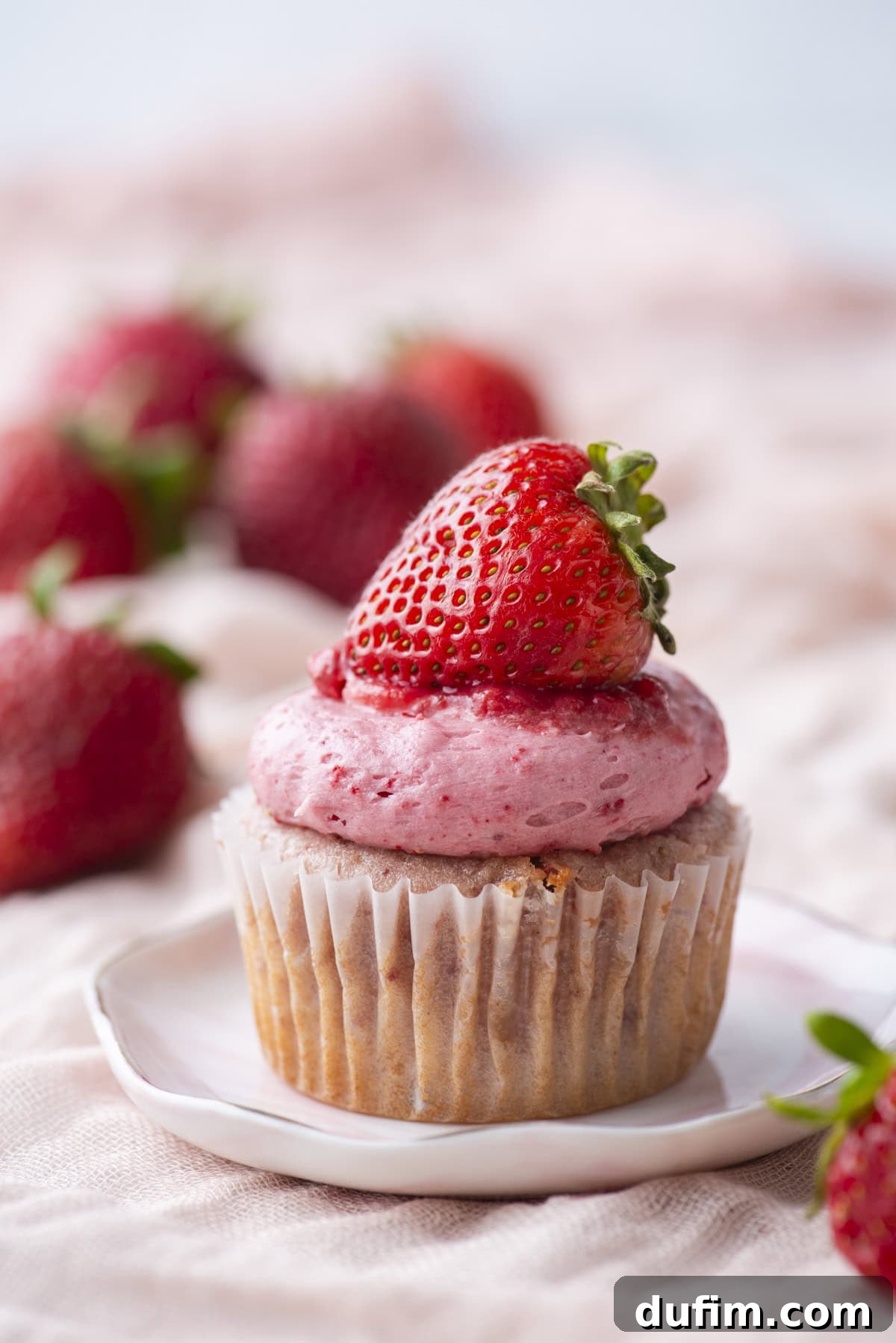 A cupcake with pink strawberry frosting, topped with a fresh whole strawberry, sits on a small white plate. Blurred strawberries are visible in the background.