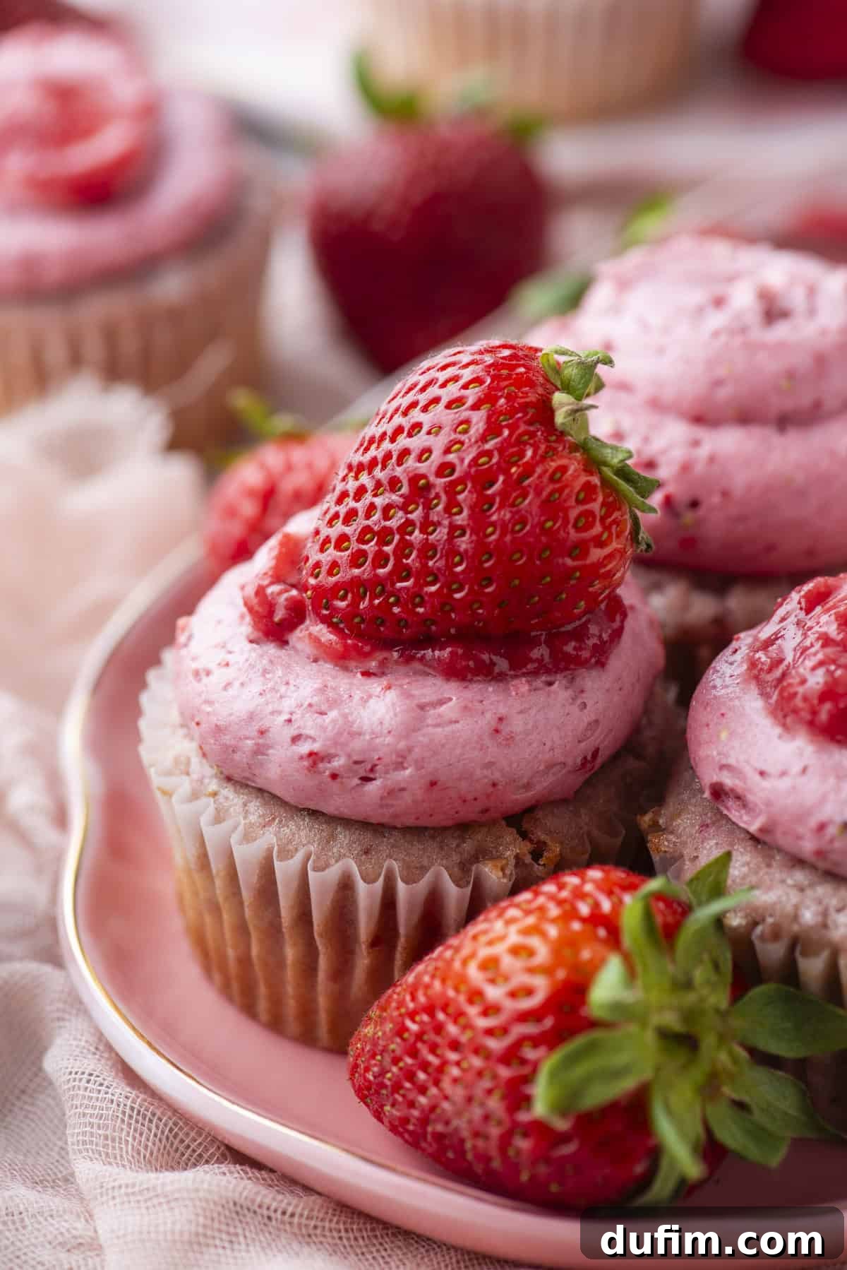 Close-up of cupcakes topped with pink strawberry frosting and fresh strawberries, arranged on a pink plate with whole strawberries around them. The background is softly blurred.