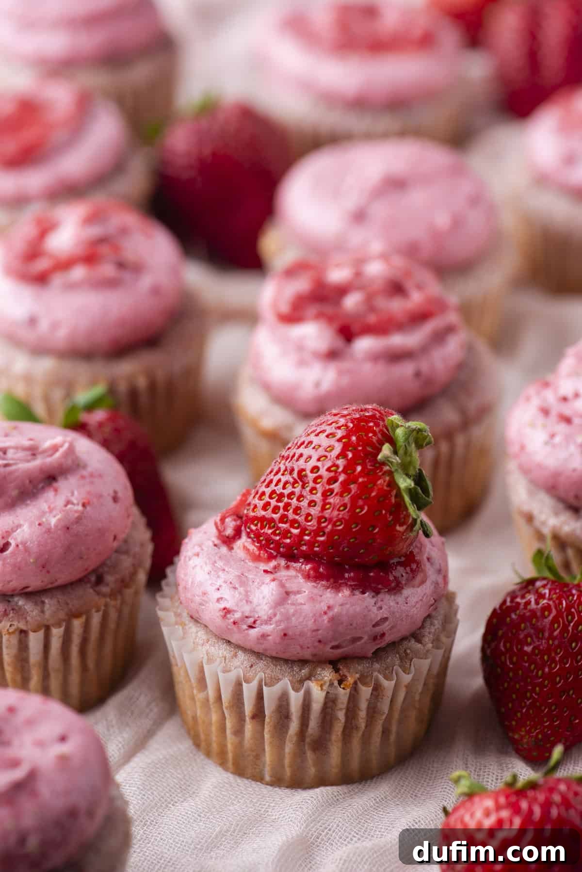 Cupcakes topped with pink strawberry frosting are arranged in rows. Some cupcakes are decorated with fresh strawberries and a drizzle of strawberry sauce. Whole strawberries are scattered among the cupcakes.