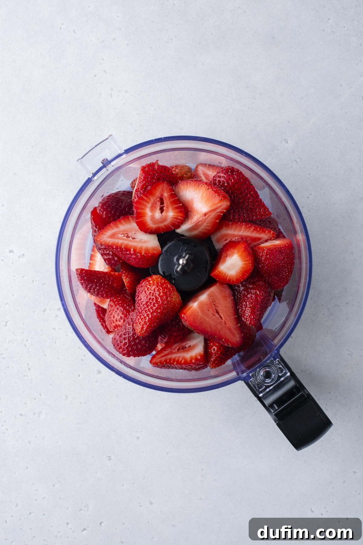 Top view of a food processor filled with fresh, halved strawberries on a light gray surface.