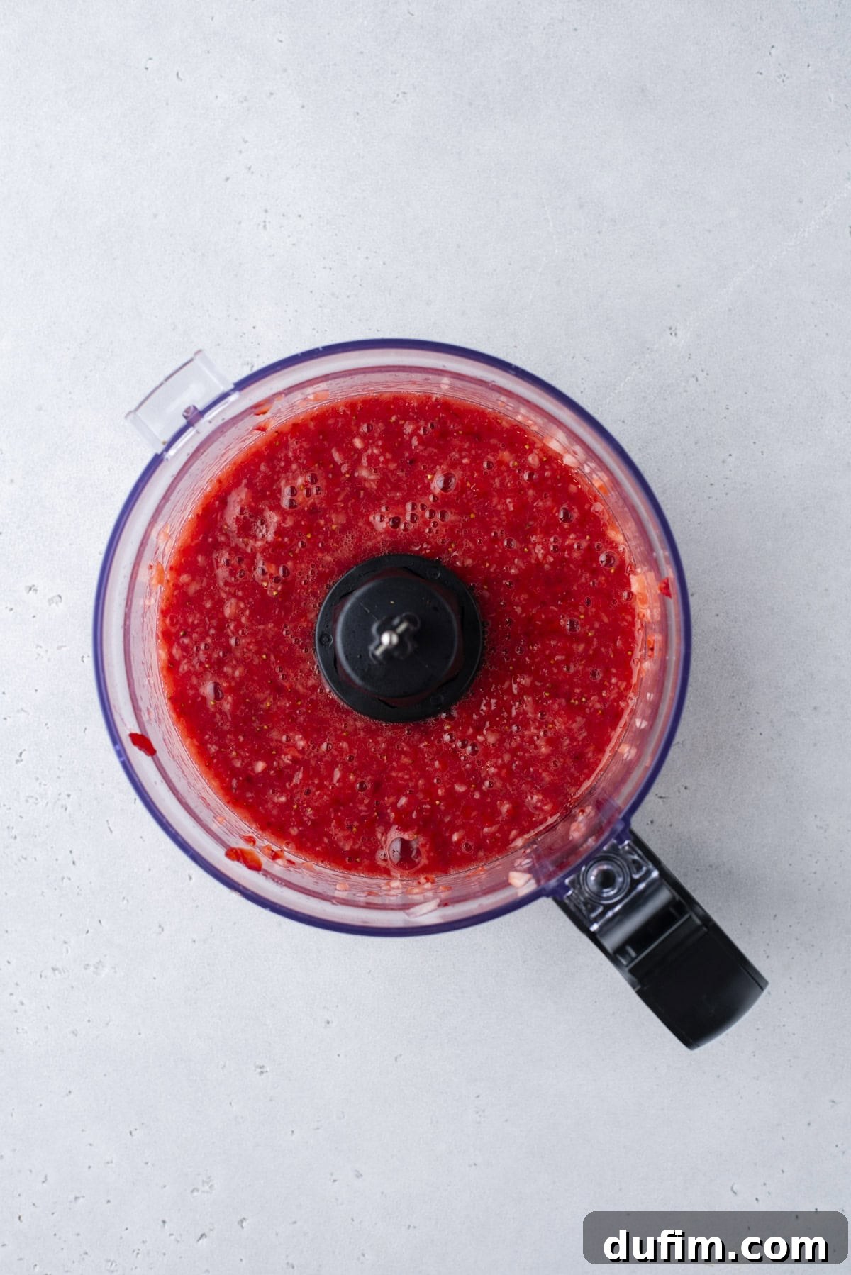 A food processor filled with a bright red, blended mixture, likely fruit puree or sauce, sits on a light gray countertop.