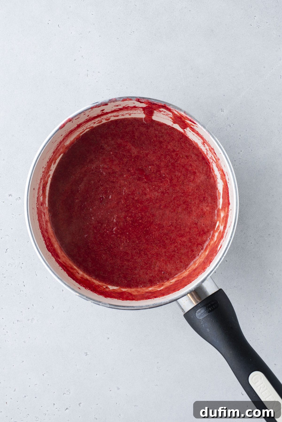 A saucepan with a red, smooth fruit sauce or puree inside, resting on a light gray surface. The sauce coats the sides of the pan, and the handle is black with a brand name visible.