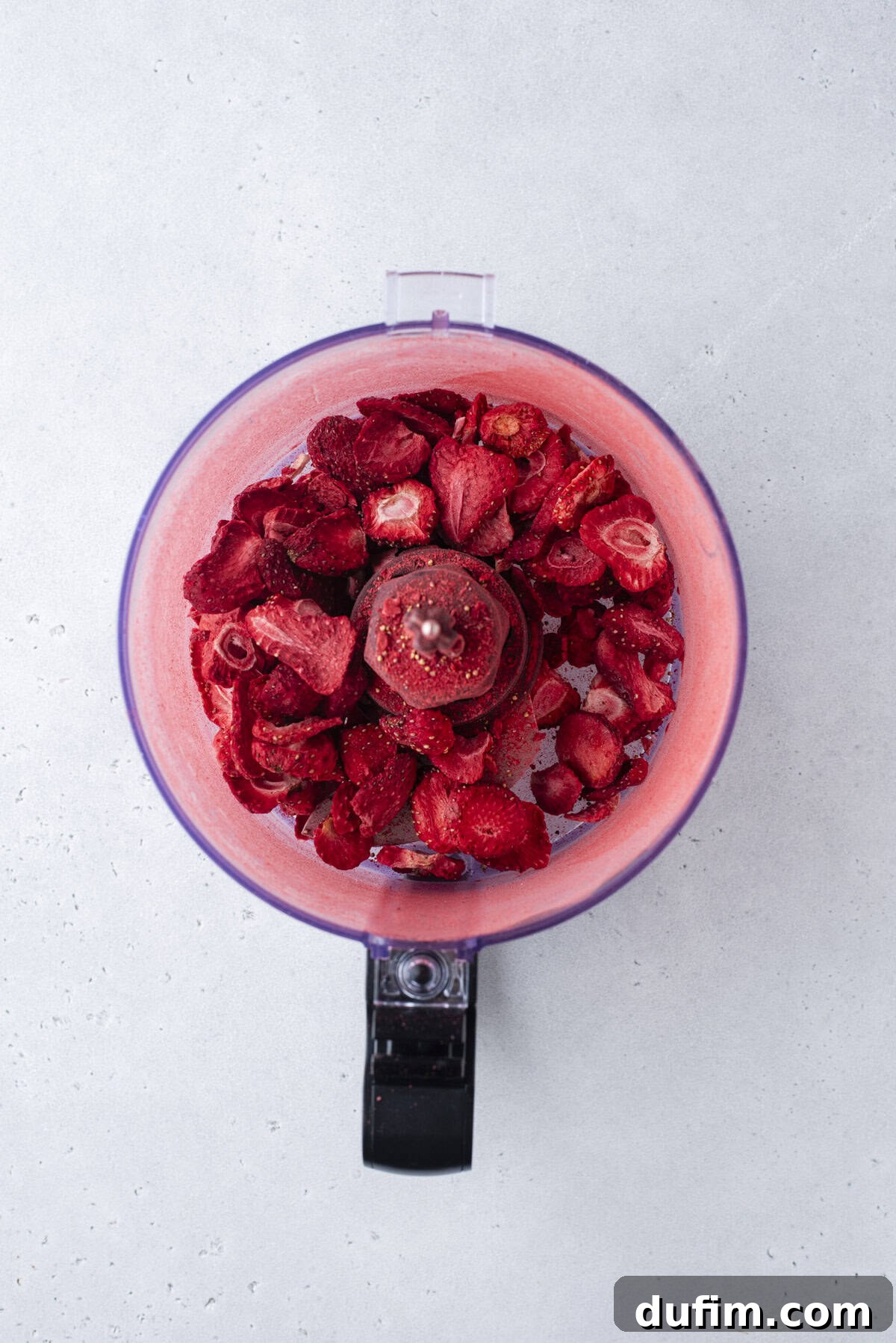 A food processor bowl filled with freeze-dried strawberries, viewed from above on a light gray surface.