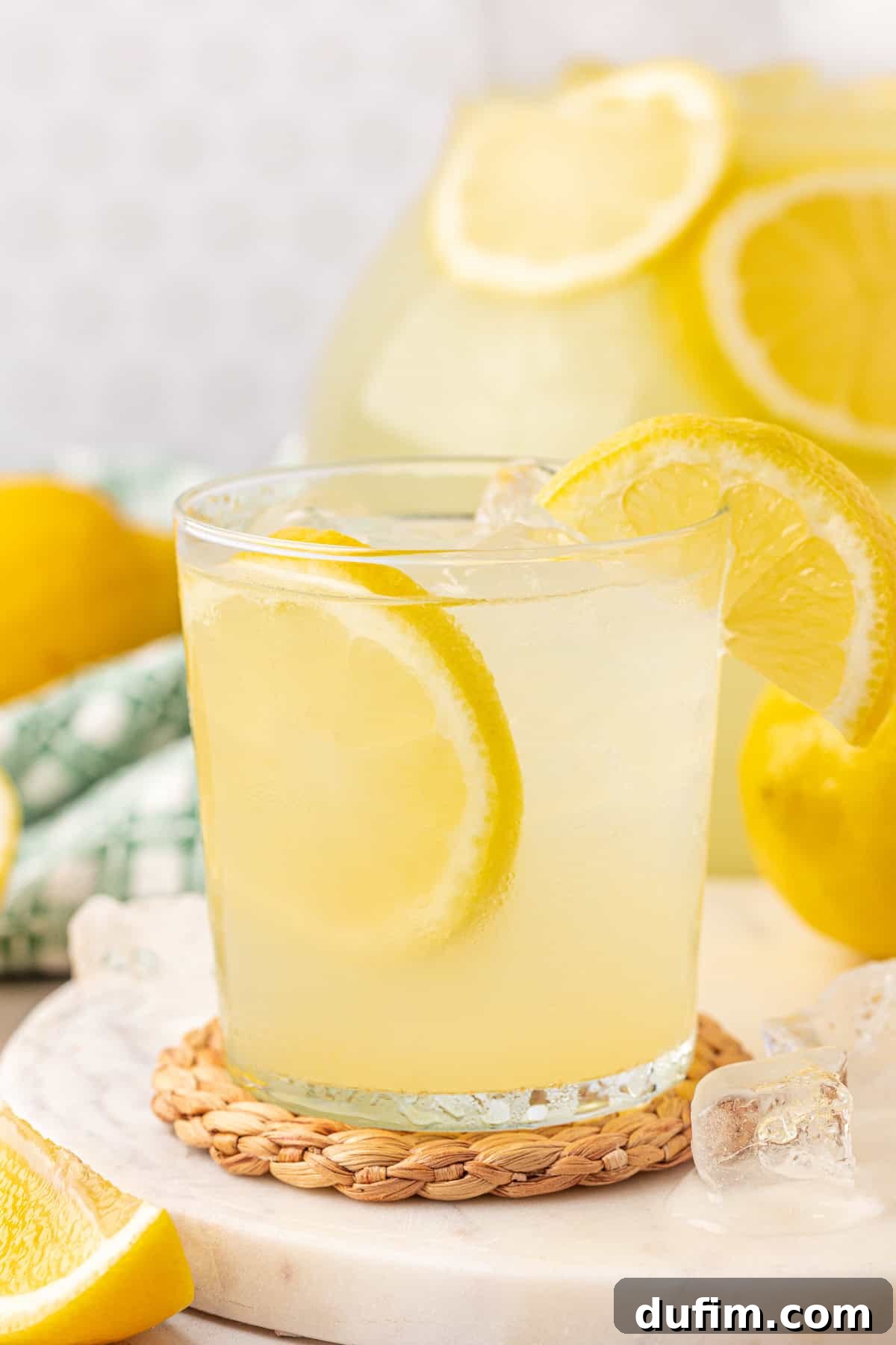Homemade Lemonade Perfection 2 A glass of lemonade with ice and a lemon slice garnish sits on a woven coaster, with a pitcher of lemonade and additional lemon slices in the background.