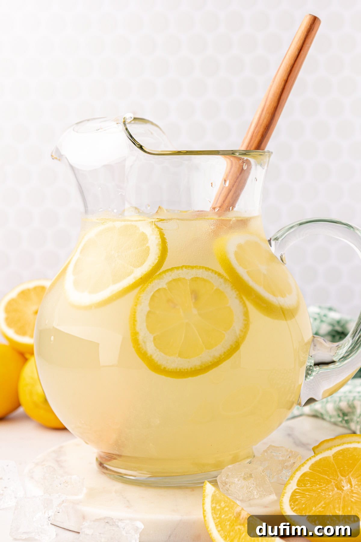 Homemade Lemonade Perfection 11 A glass pitcher filled with lemonade and lemon slices, with a wooden spoon inside. Ice cubes and lemon wedges are scattered around the pitcher on a white surface.