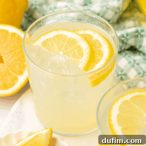 A glass of lemonade with ice and lemon slices, placed on a white surface next to a halved lemon and a patterned green and white cloth in the background.