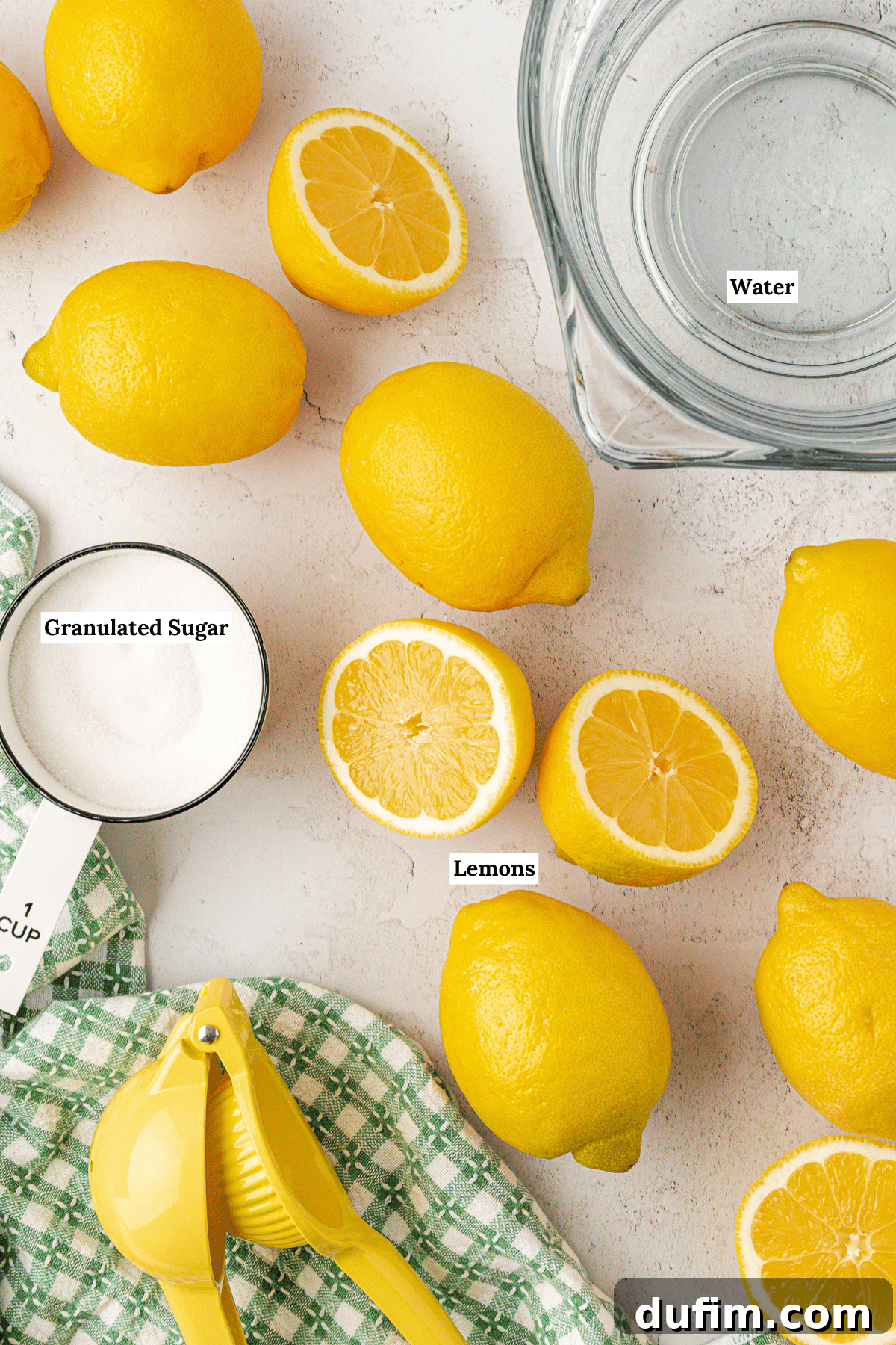 Overhead view of whole and halved lemons, a bowl of water, a measuring cup of granulated sugar, a yellow citrus juicer, and a green checkered cloth on a white surface. Some items are labeled.
