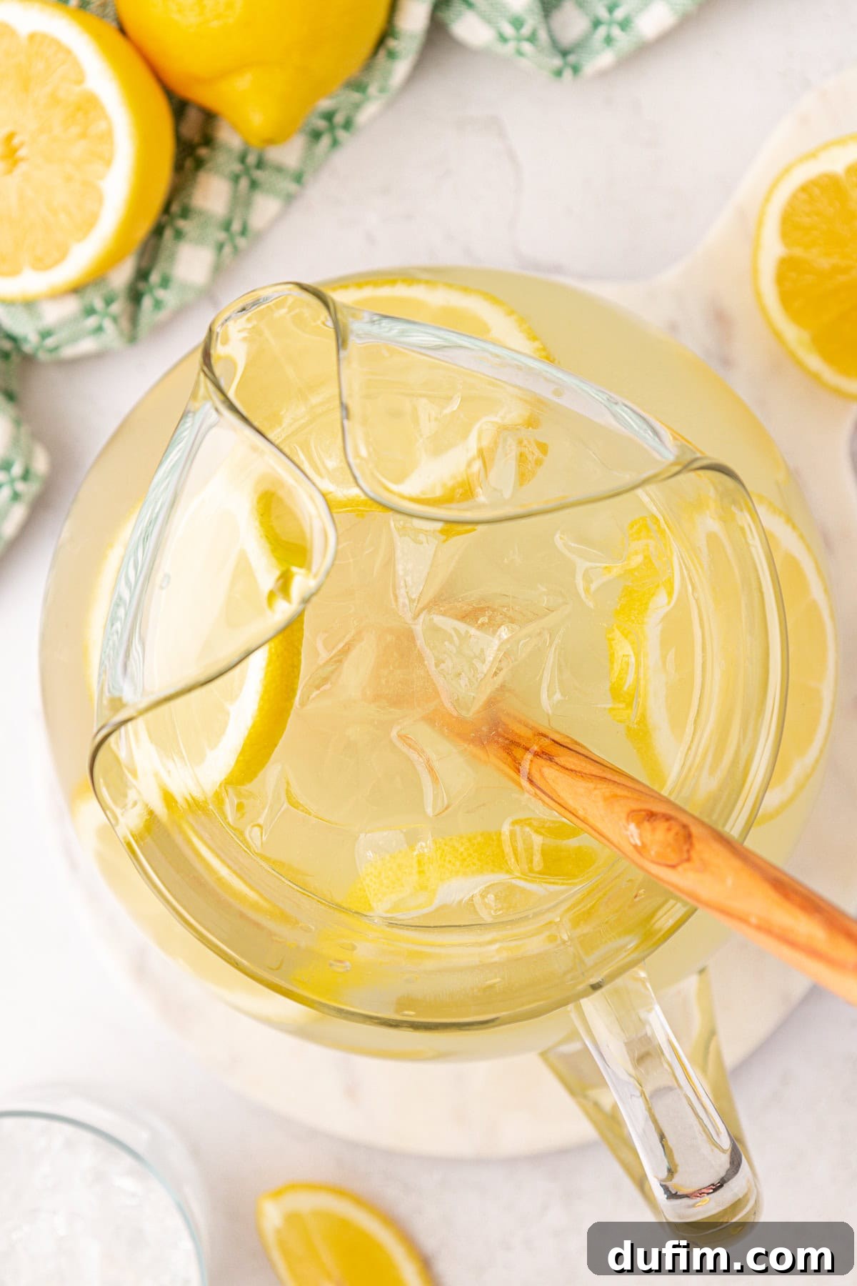 Homemade Lemonade Perfection 4 A top view of a glass pitcher filled with iced lemonade and lemon slices, being stirred with a wooden spoon. Fresh lemon halves and a green checkered cloth are visible in the background.