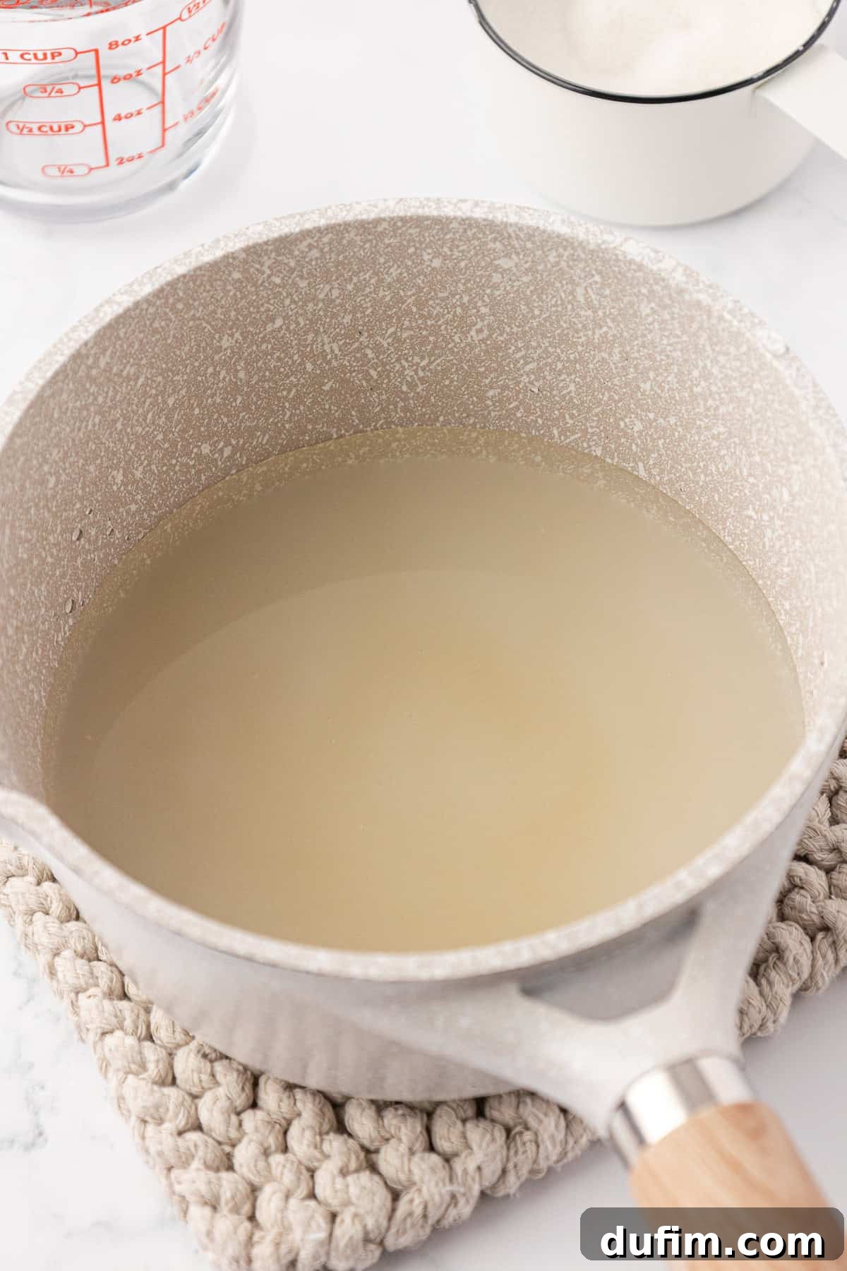 Homemade Lemonade Perfection 5 A saucepan filled with a cloudy liquid sits on a knitted mat, with a measuring cup of water and a small white pot of sugar in the background on a marble surface.