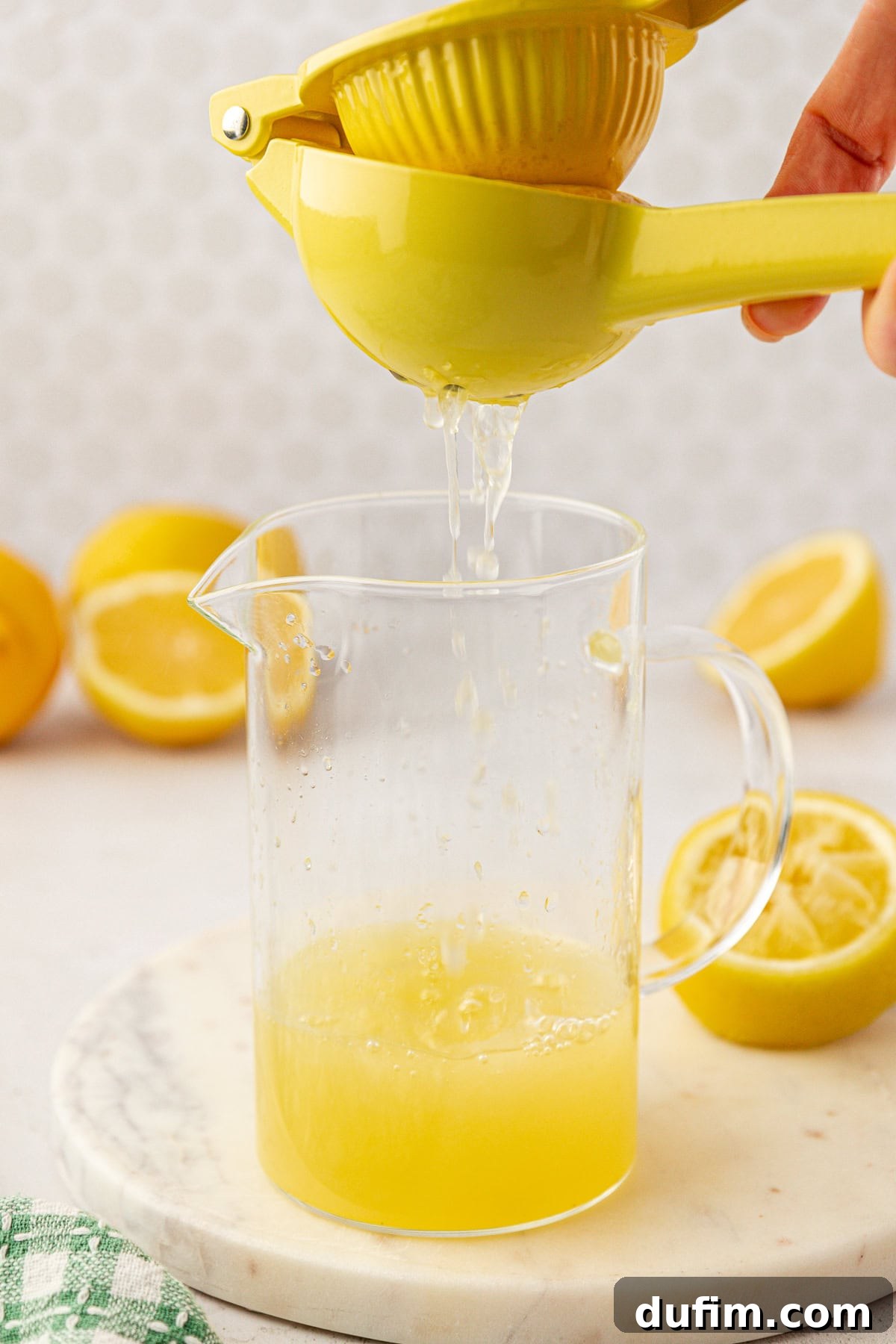 Homemade Lemonade Perfection 7 A hand squeezes fresh lemon juice with a yellow citrus press over a clear glass pitcher, surrounded by halved lemons on a marble surface.