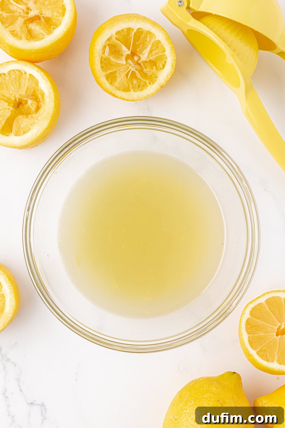 Homemade Lemonade Perfection 8 A glass bowl filled with lemon juice is surrounded by halved and whole lemons, with a yellow citrus juicer nearby on a white surface.