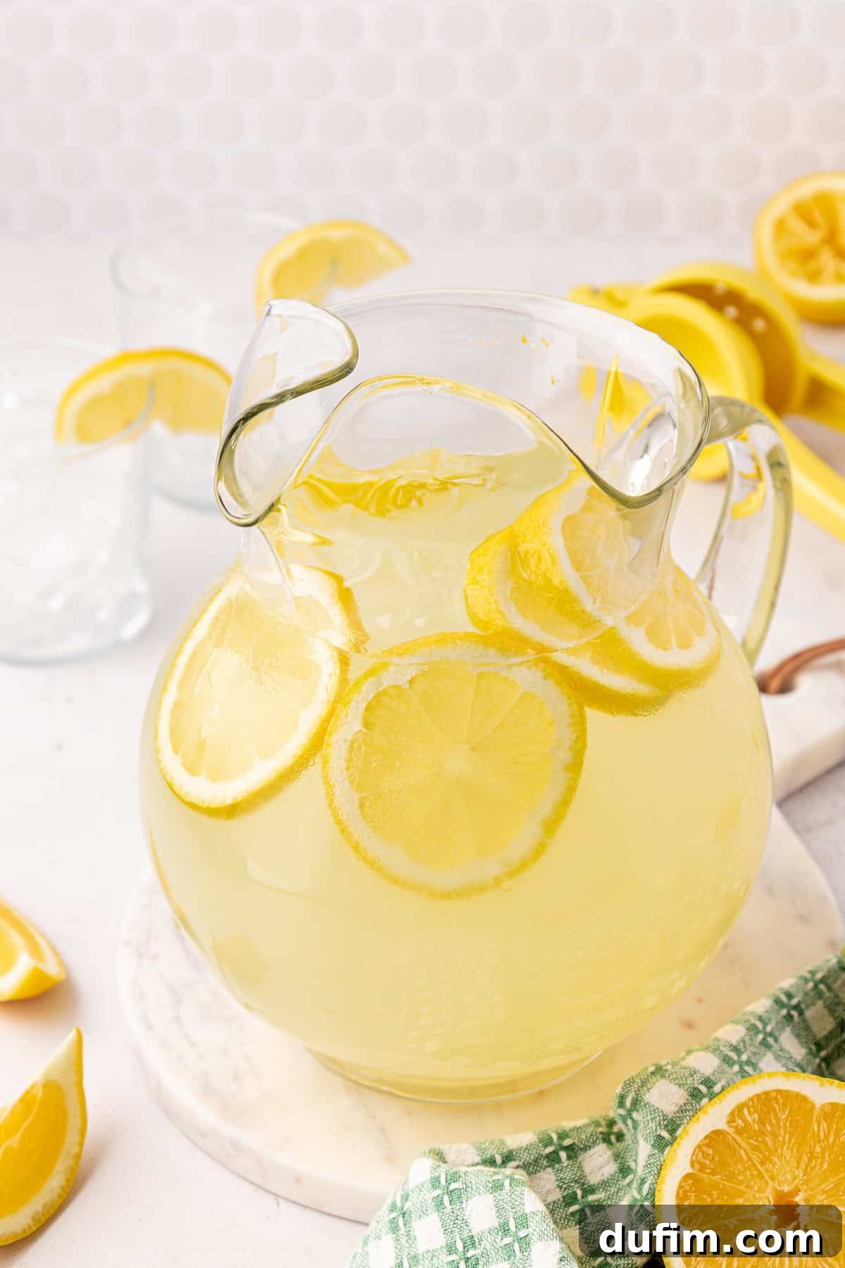 Homemade Lemonade Perfection 9 A glass pitcher of lemonade with floating lemon slices sits on a marble surface. Lemon wedges, a green checkered towel, and a yellow citrus squeezer are nearby.