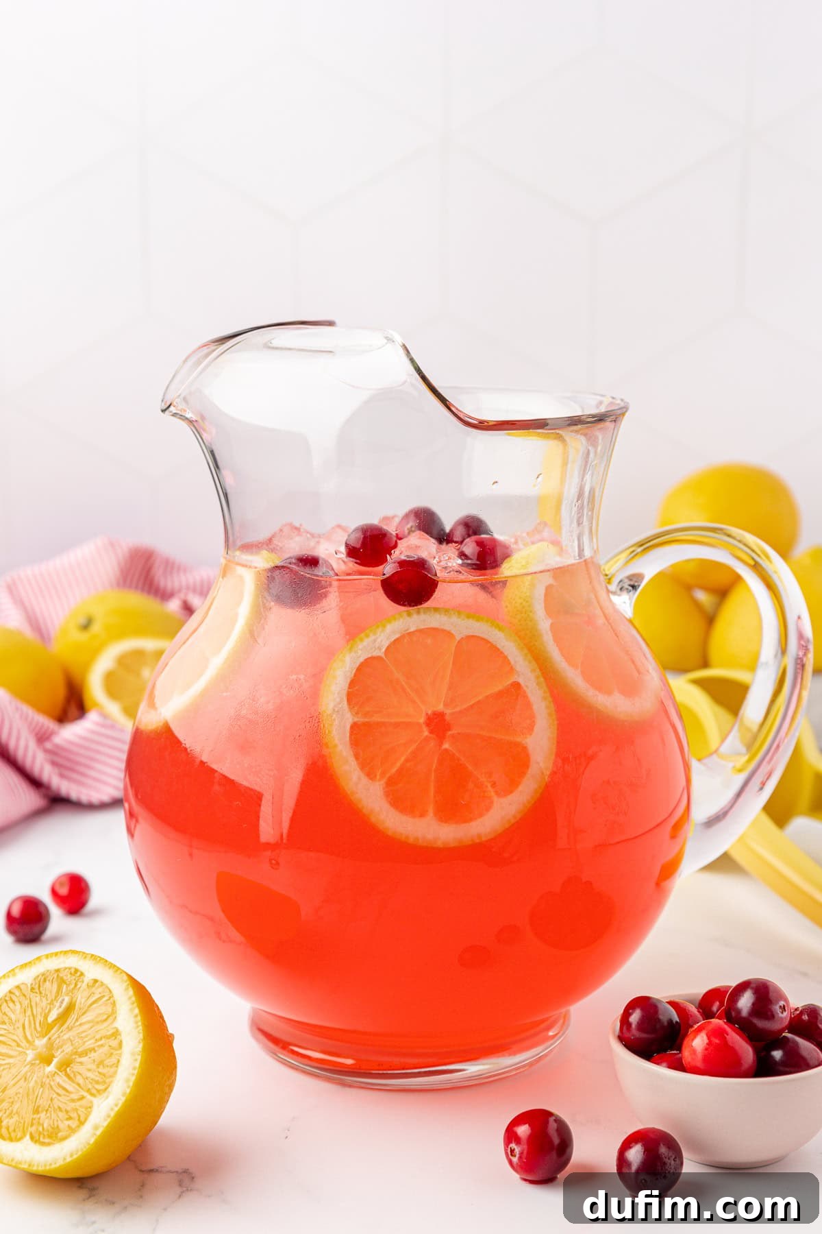 Your Guide to Perfect Homemade Pink Lemonade 2 A clear glass pitcher filled with pink lemonade, garnished with lemon slices and cranberries, sits on a white counter near a small bowl of cranberries and fresh lemons.