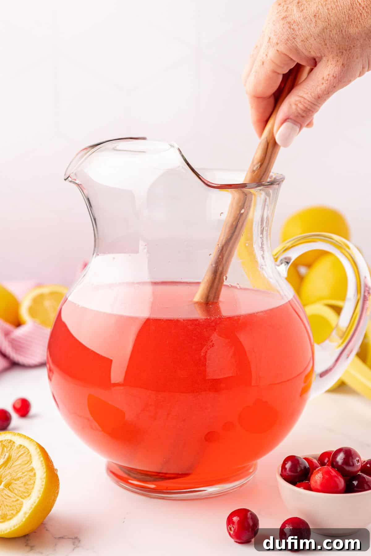 Your Guide to Perfect Homemade Pink Lemonade 11 A hand stirs a pitcher of bright pink punch with a wooden spoon. The pitcher is on a white surface, surrounded by fresh lemons, cranberries, and a small bowl of cranberries.