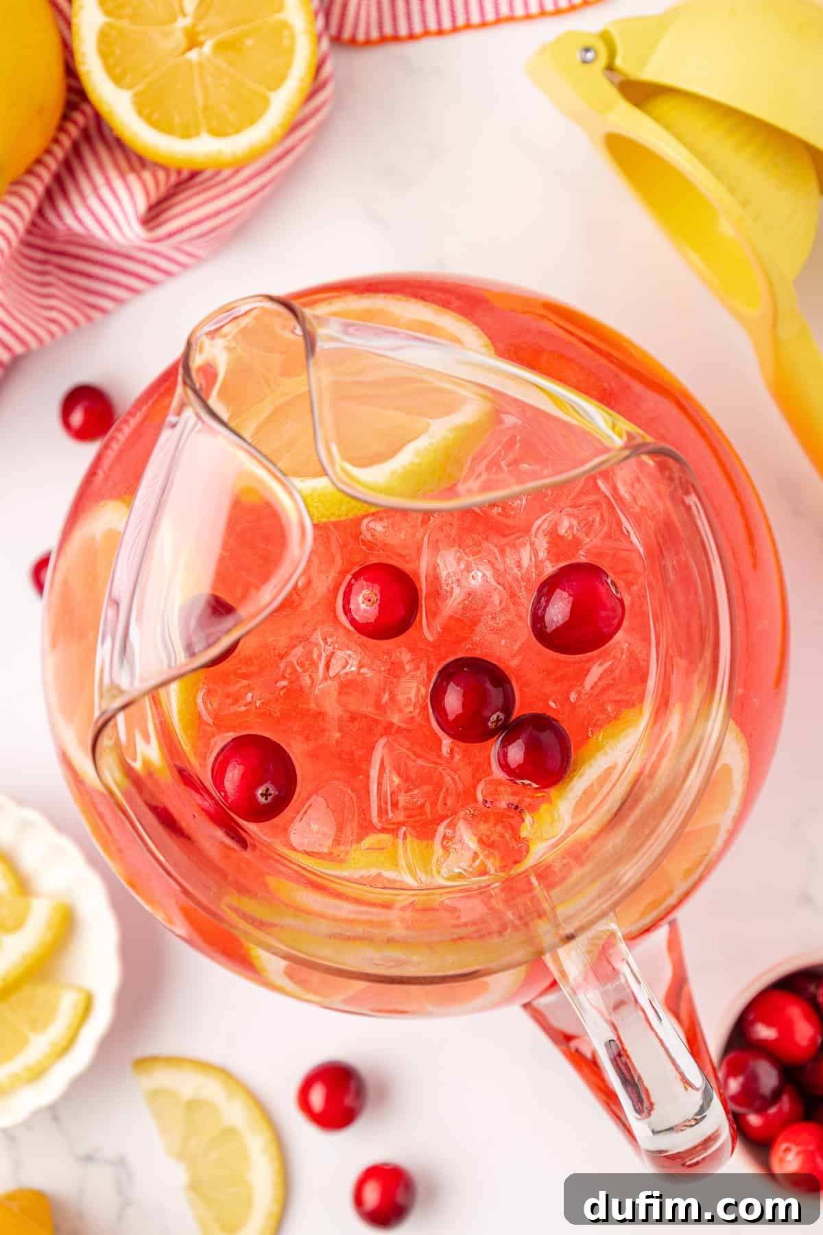 Your Guide to Perfect Homemade Pink Lemonade 13 A glass pitcher filled with pink lemonade, ice, lemon slices, and cranberries sits on a white surface surrounded by fresh lemon wedges, cranberries, a citrus juicer, and a red-striped cloth.