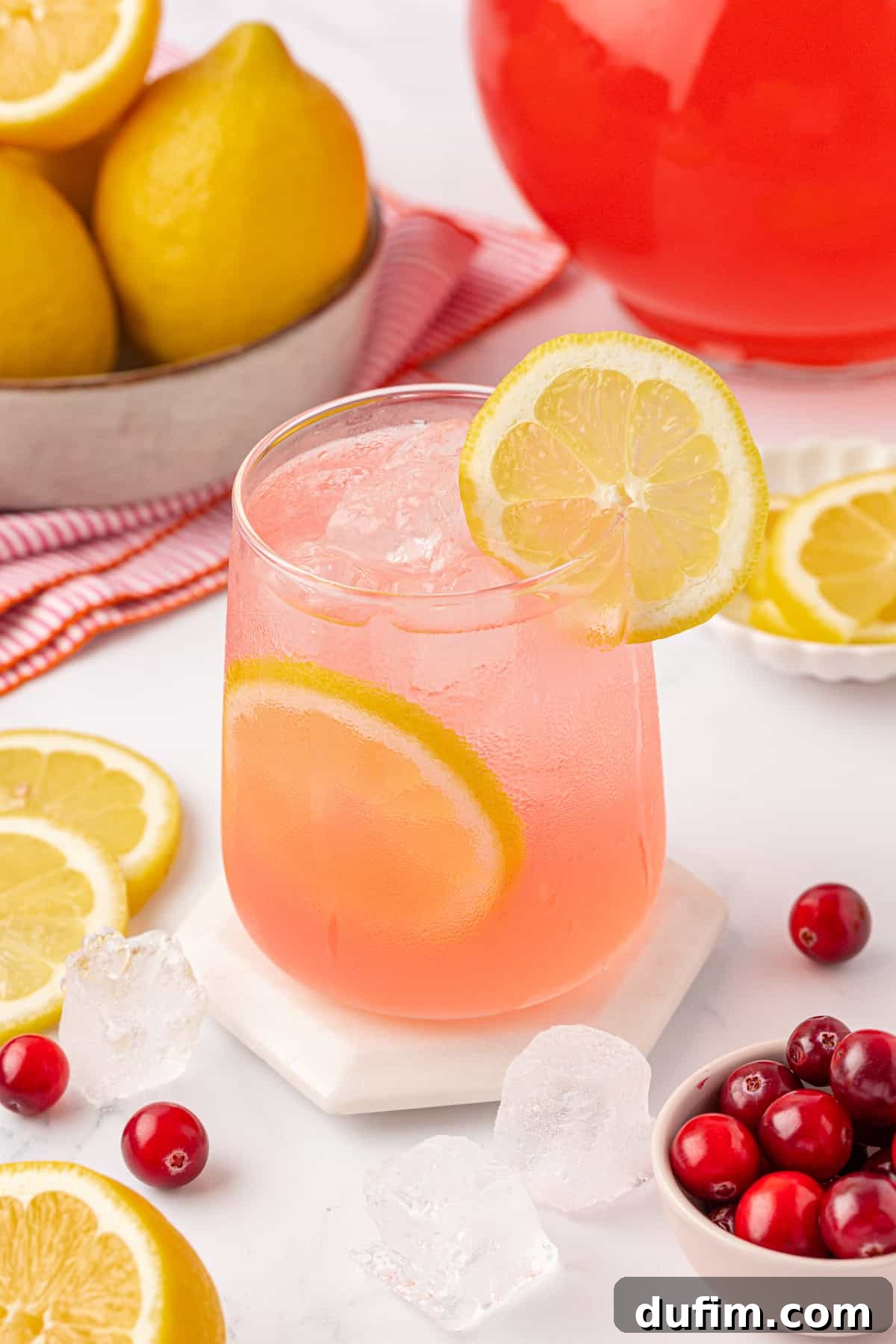 Your Guide to Perfect Homemade Pink Lemonade 14 A glass of pink lemonade with ice and lemon slices, garnished with a lemon wheel, sits on a coaster. Surrounding the glass are fresh lemons, cranberries, ice cubes, and a red pitcher.