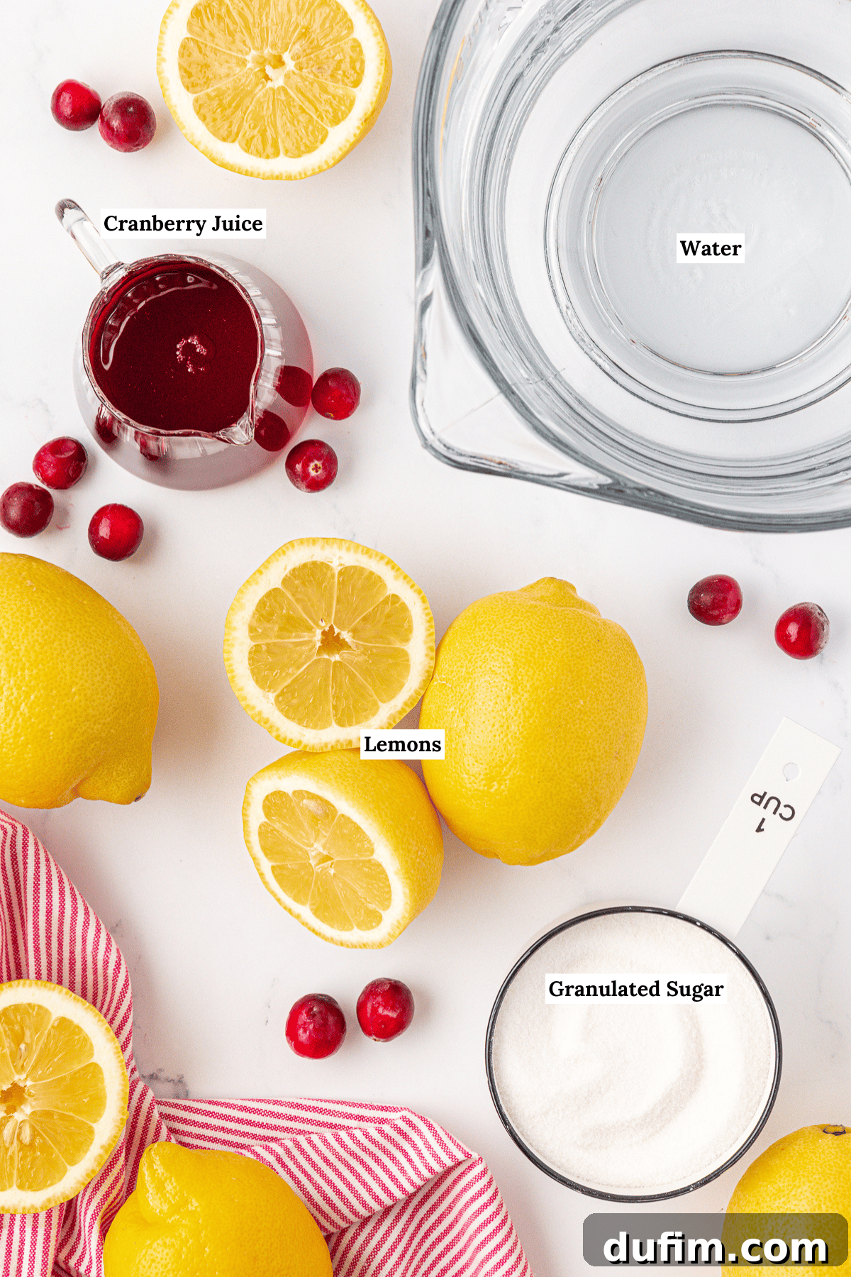 Your Guide to Perfect Homemade Pink Lemonade 4 Top-down view of ingredients for cranberry lemonade: a glass of water, cranberry juice, whole and halved lemons, granulated sugar in a measuring cup, loose cranberries, and a red-striped cloth on a white surface. Labels identify each item.