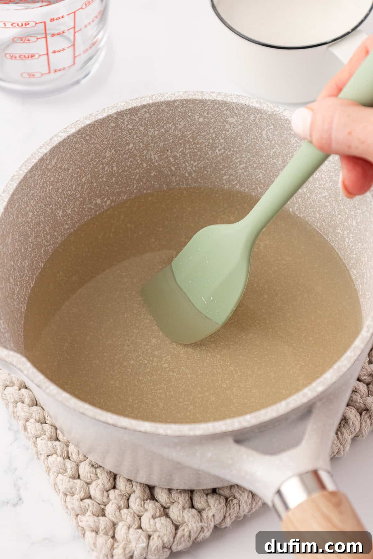 Your Guide to Perfect Homemade Pink Lemonade 7 A hand stirs a clear, light-colored liquid in a beige saucepan using a green silicone spatula; the pot sits on a woven trivet, with a measuring cup and bowl nearby.