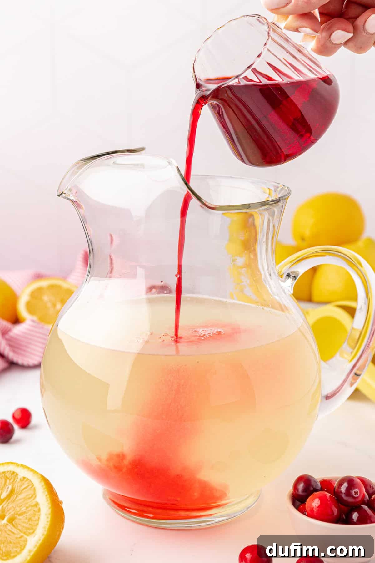 Your Guide to Perfect Homemade Pink Lemonade 10 A hand pours red liquid from a small glass pitcher into a large clear pitcher of lemonade. Lemons and cranberries are scattered around on a white surface.