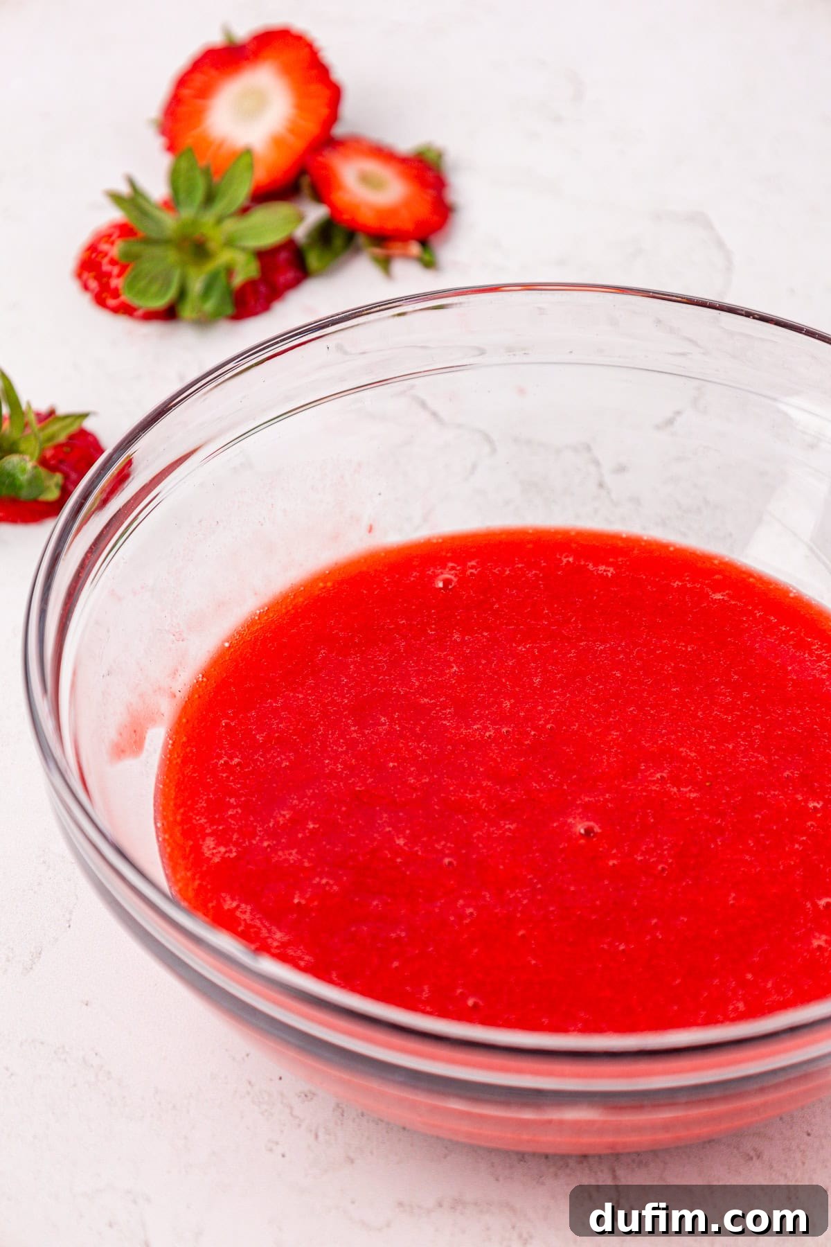 A clear glass bowl filled with vibrant, bright red strawberry puree sits on a light surface. Fresh strawberry slices and whole strawberries are artfully arranged in the background.
