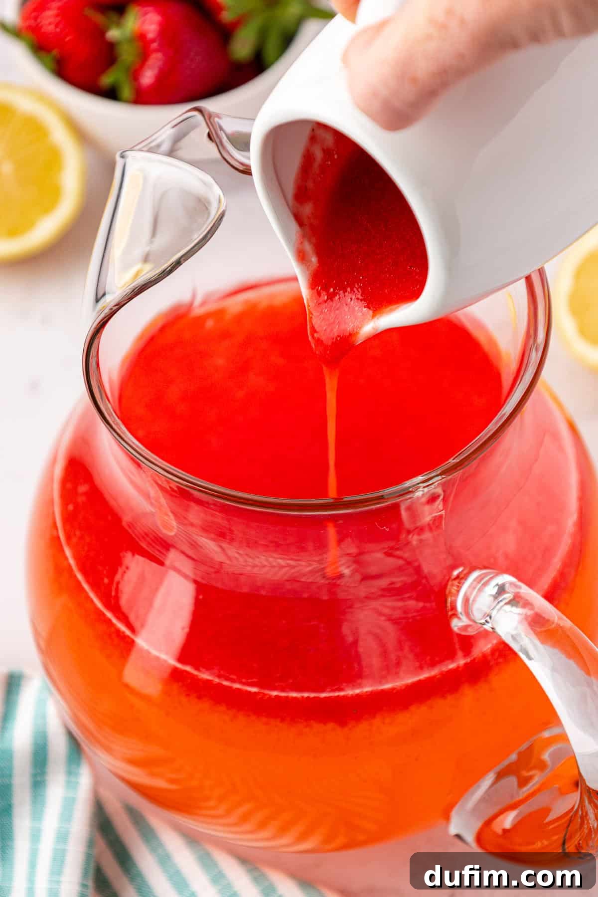 A hand gracefully pours bright red strawberry puree from a white pitcher into a clear glass jug already containing an orange-red liquid, indicating the final mixing stage. Fresh strawberries, lemon slices, and a striped towel are arranged nearby.