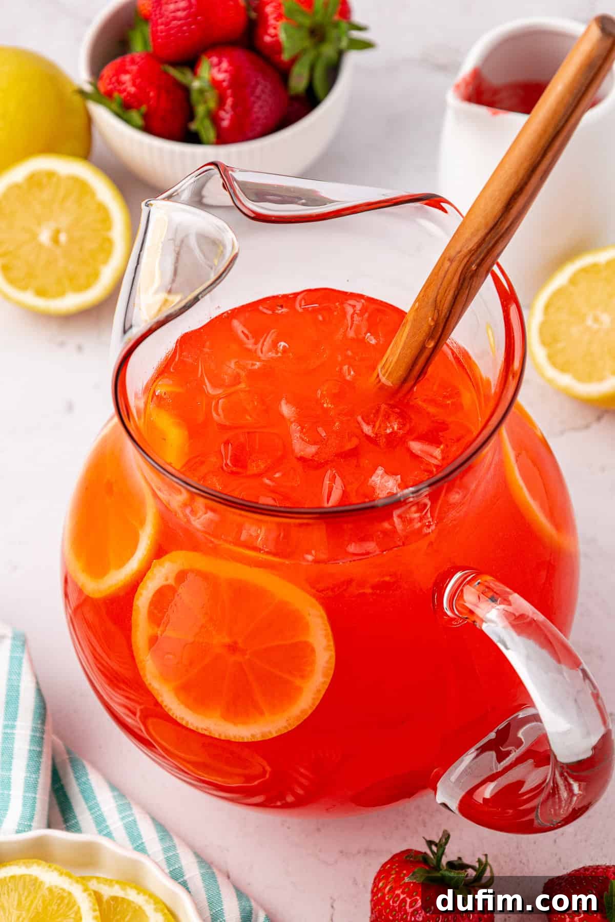 A elegant glass pitcher brimming with iced strawberry lemonade, adorned with vibrant lemon slices, sits on a rustic table. Fresh strawberries and lemon halves are scattered around, completing a picturesque summer display. A wooden spoon rests invitingly inside the pitcher.