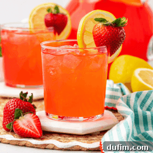 A glass of bright red strawberry lemonade with ice, garnished with a lemon slice and strawberry. Another drink, strawberries, lemons, and a striped cloth are in the background on a woven mat.