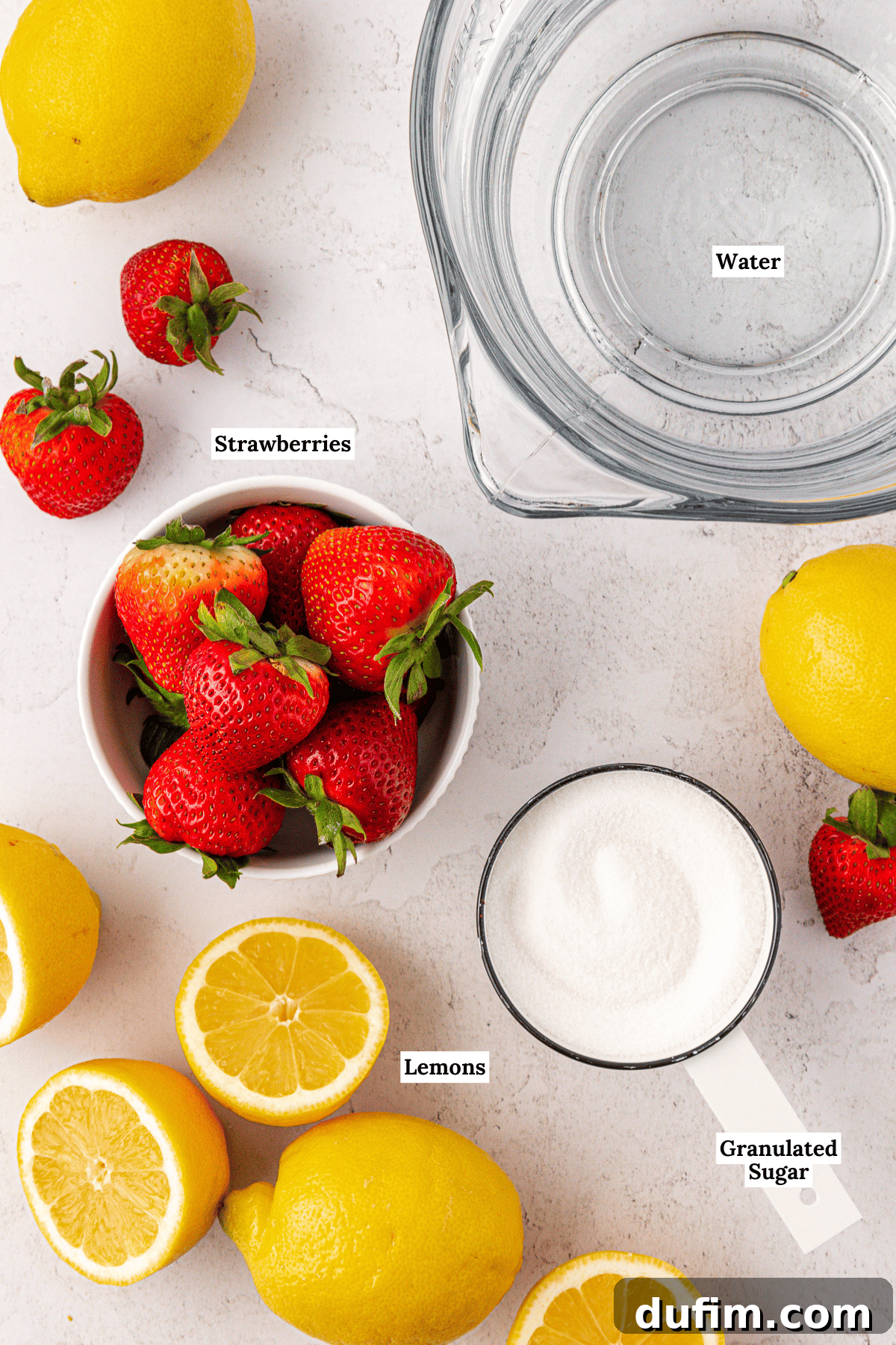 A top-down view of fresh ingredients on a white surface: vibrant strawberries, halved and whole lemons, granulated sugar in a measuring cup, and a bowl of water. Each essential component for homemade strawberry lemonade is neatly arranged.